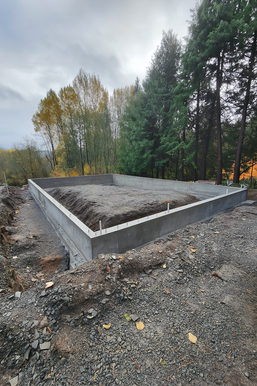 A concrete foundation for a building filled with gravel, set on a construction site surrounded by trees.