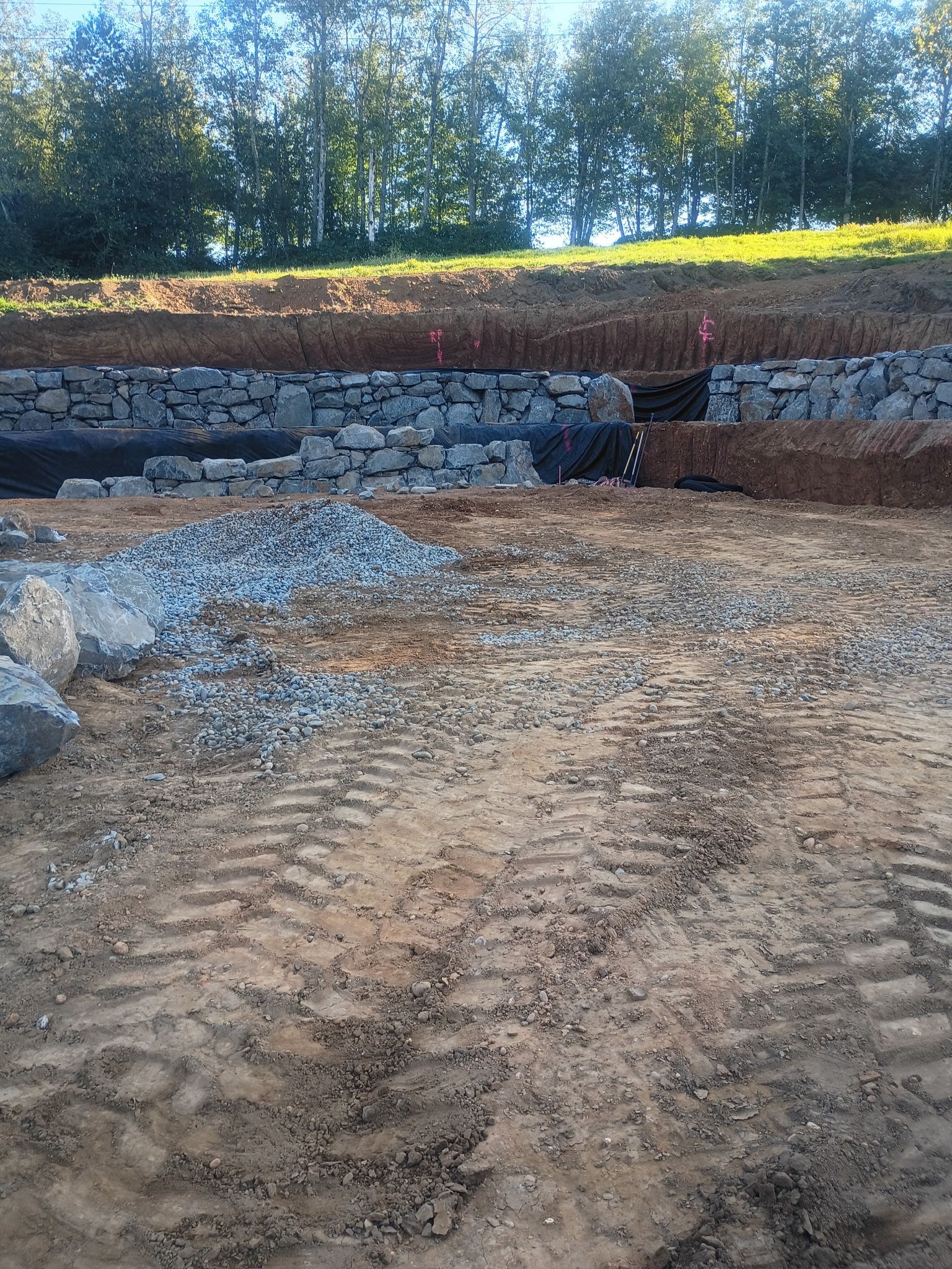 A stone retaining wall under construction at the edge of a dirt site with tire tracks, leading up to a treeline.