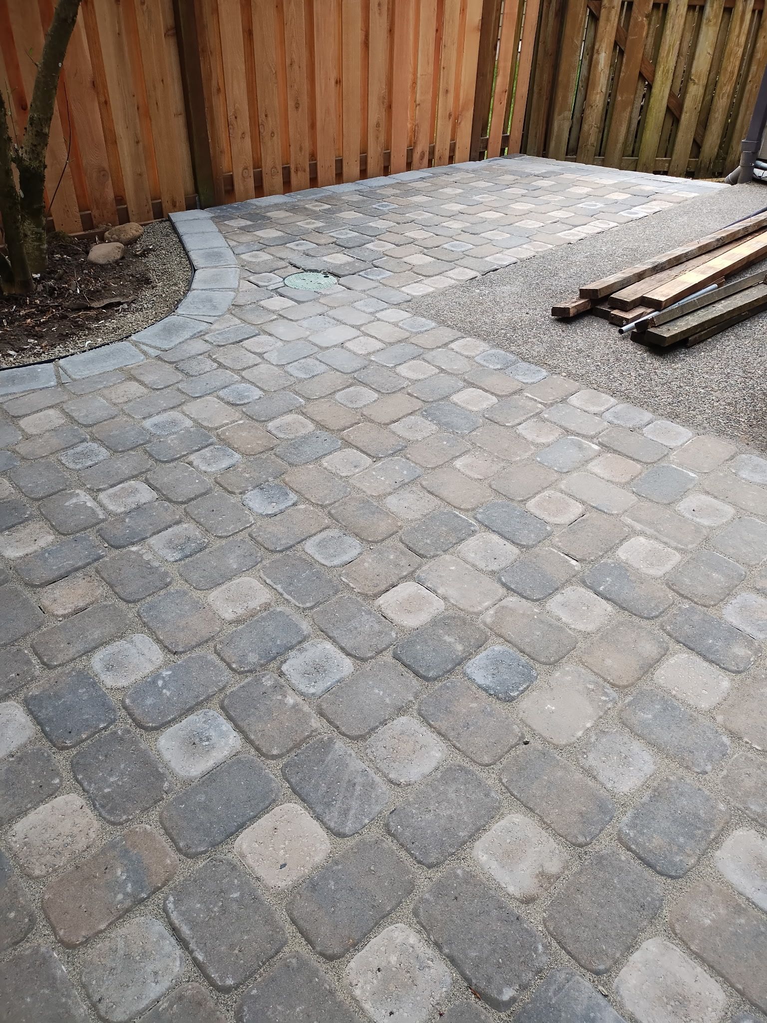 A patio made of grey, multi-sized interlocking pavers with a rounded border, situated next to a wooden fence and gravel.