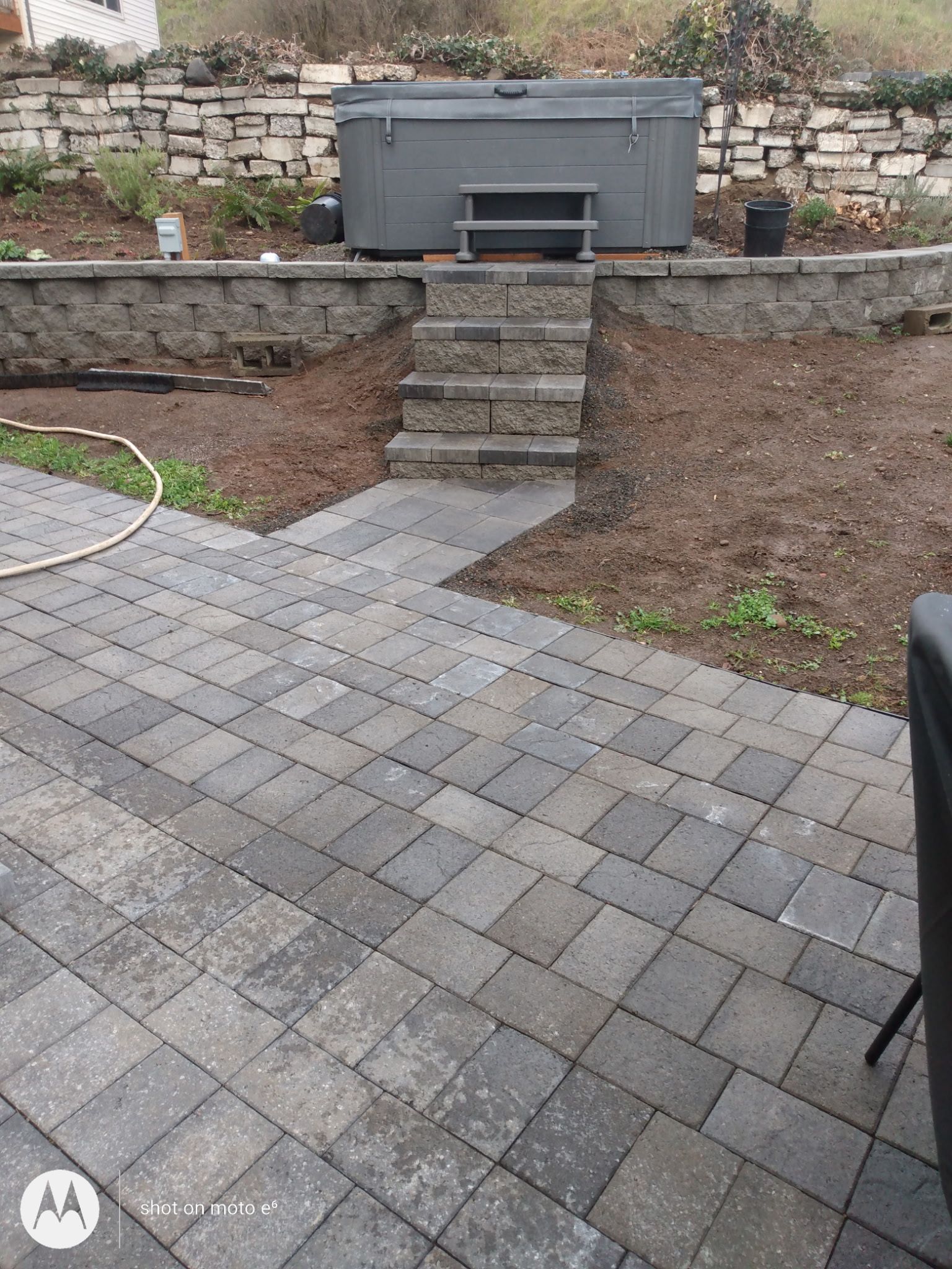 A backyard scene showing a gray hot tub on a raised stone terrace with stairs leading down to a gray paver patio.