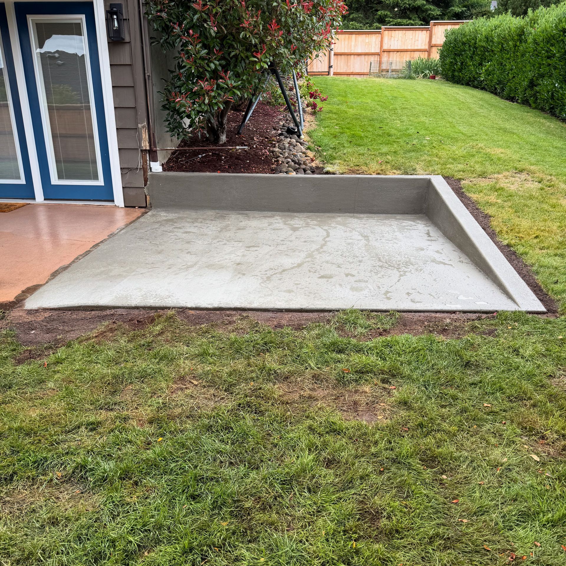 A newly poured concrete patio in a backyard, featuring a retaining wall and a sloped edge leading into the grass.