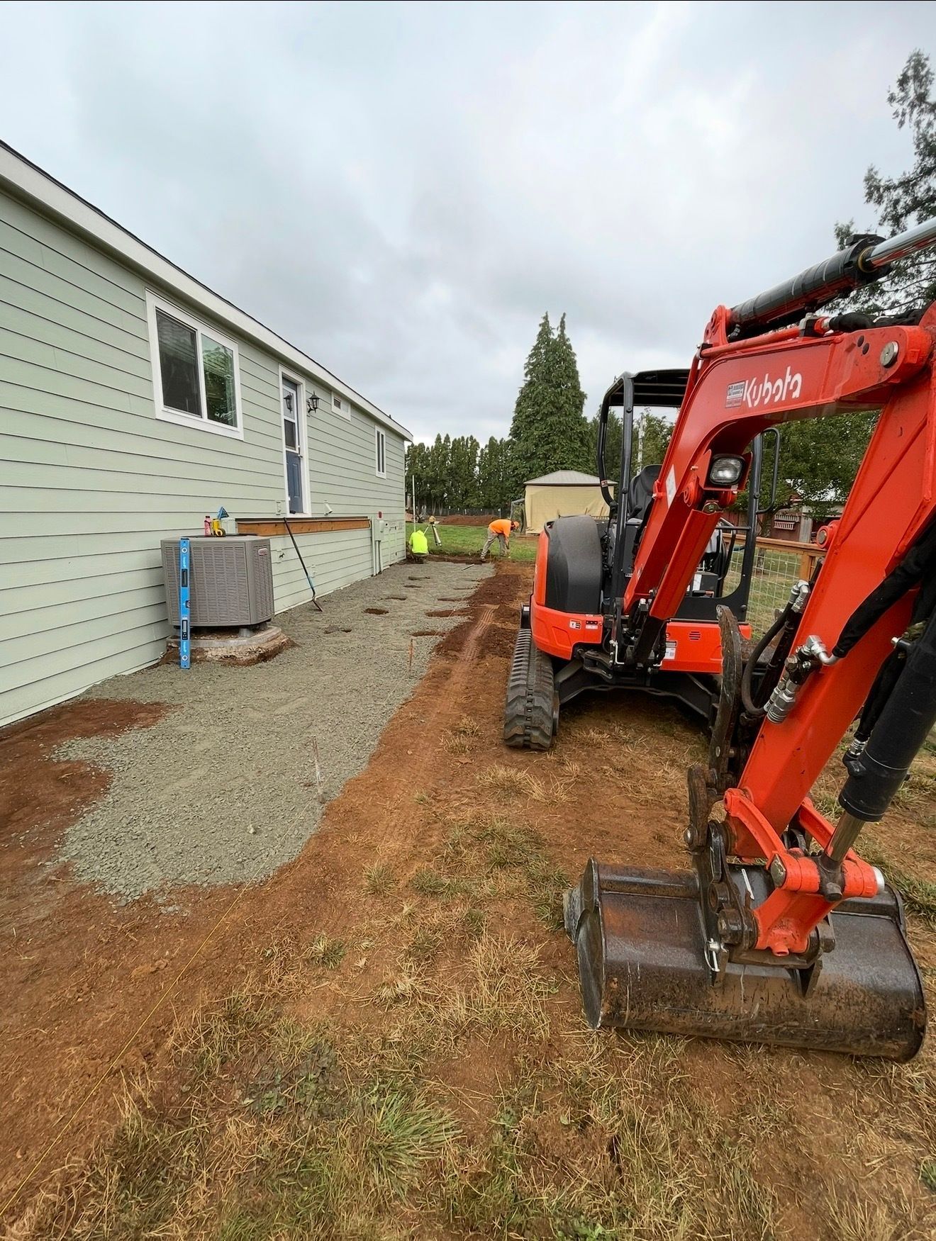 An orange mini-excavator sits on a dirt lot next to a light-green house with a gravel path during exterior construction.