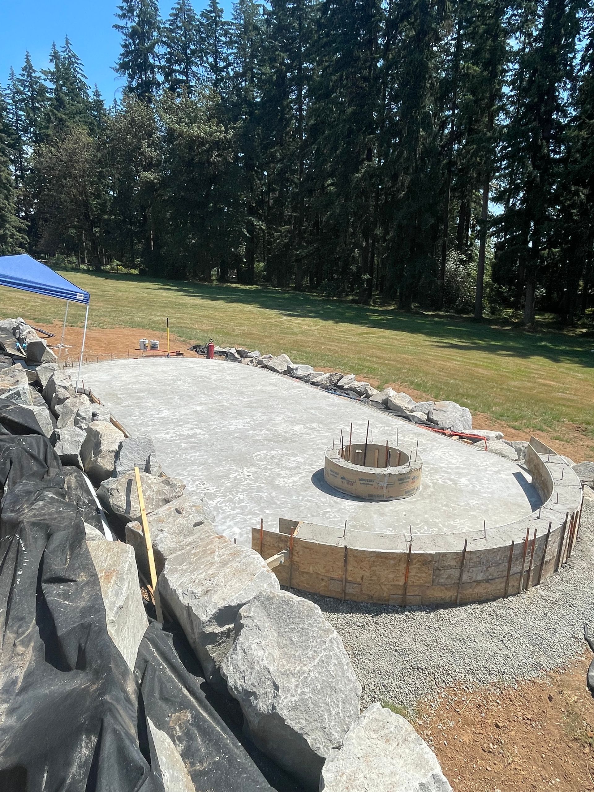 An outdoor stone fire pit under construction with a gravel base, surrounded by stone walls and a treeline in the distance.