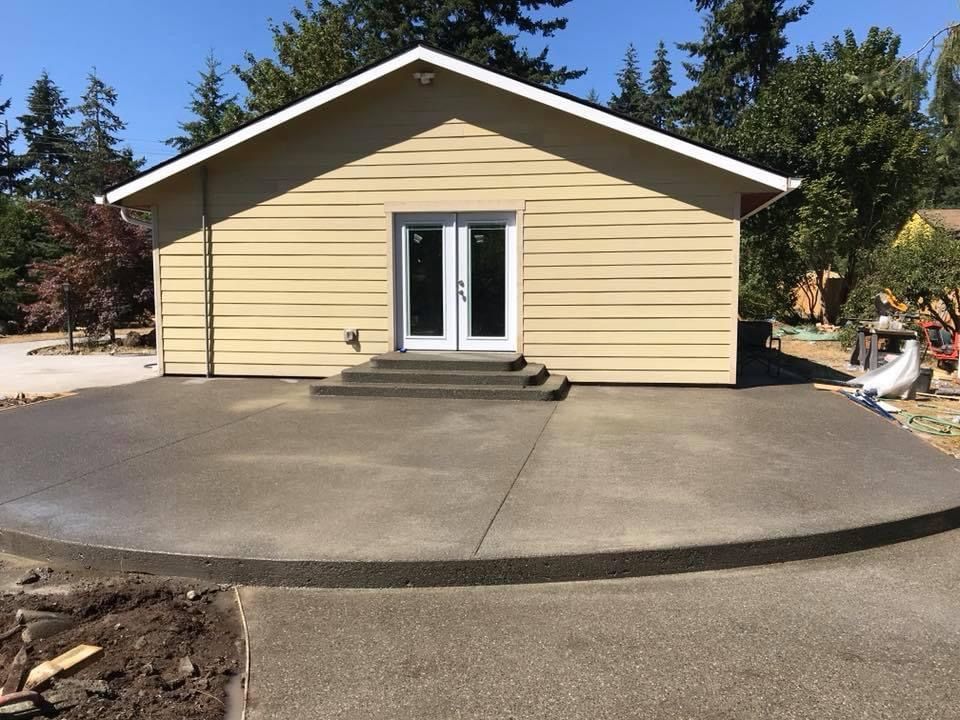 A tan building with double doors and concrete stairs, leading to a semi-circular concrete patio in a sunny outdoor setting.