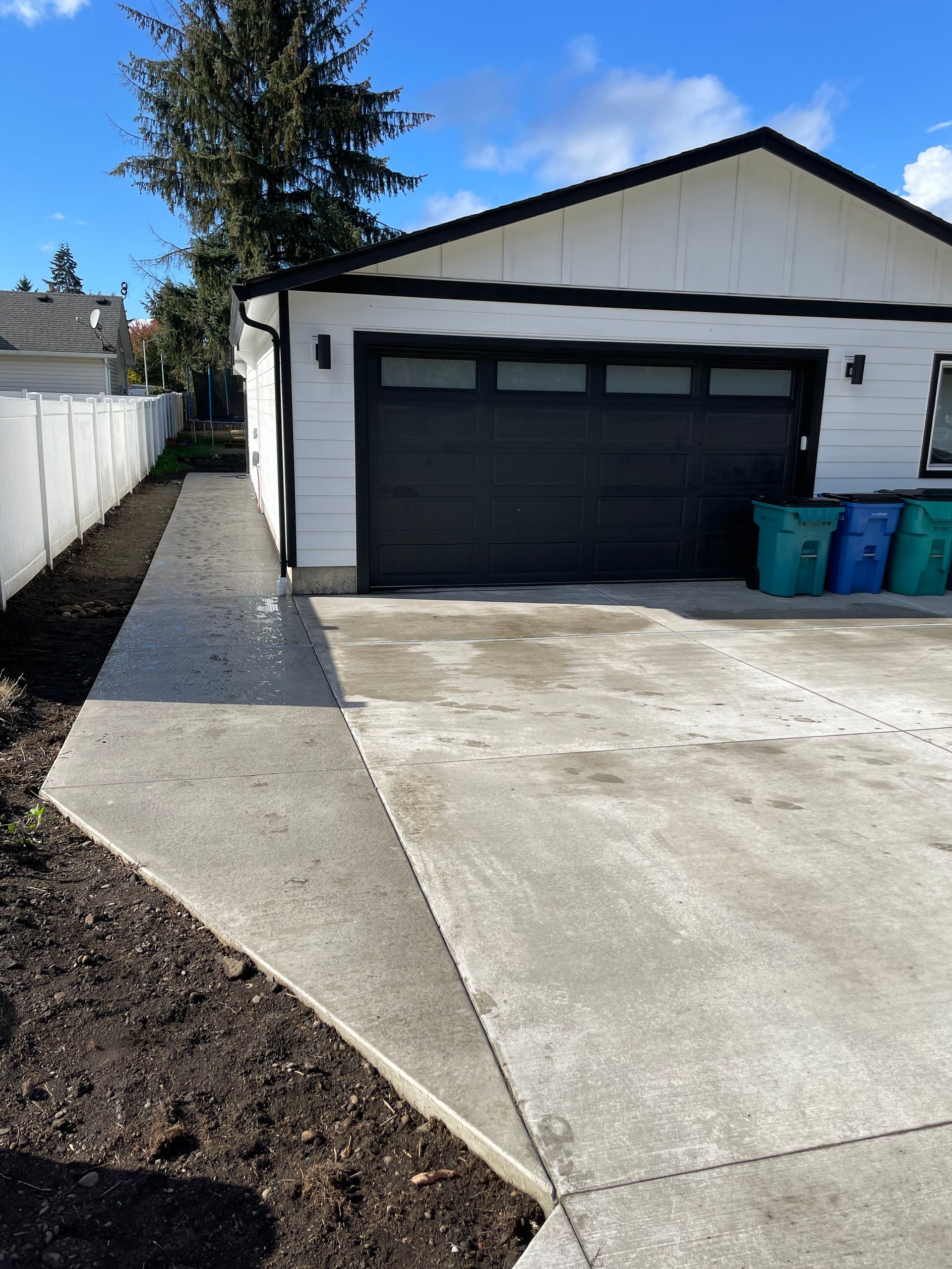 A white detached garage with a black door and concrete driveway, next to a path and a fenced area with recycling bins.