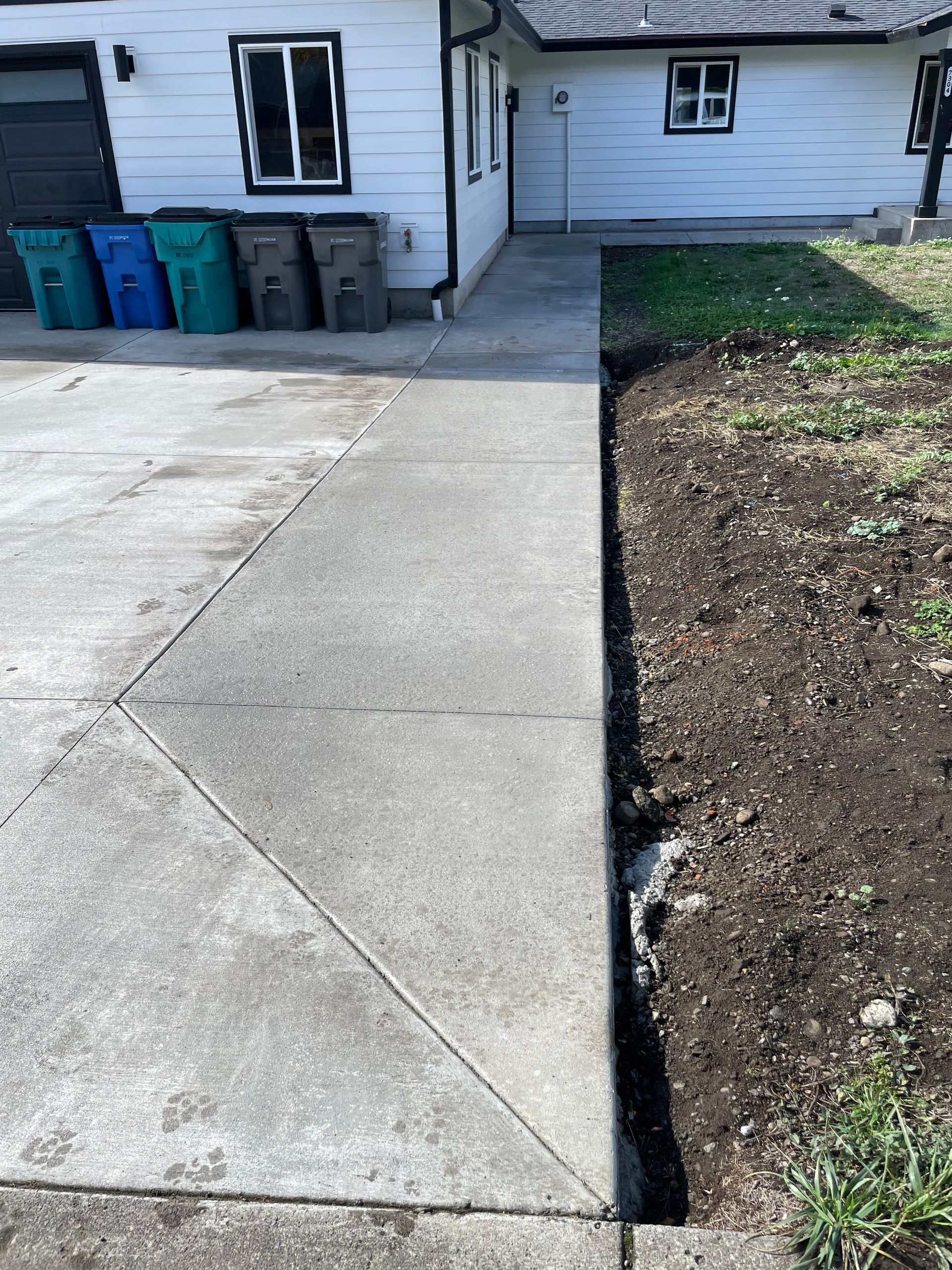 A white house with a concrete walkway leading to an entrance, next to a patch of dark soil and four plastic waste bins.