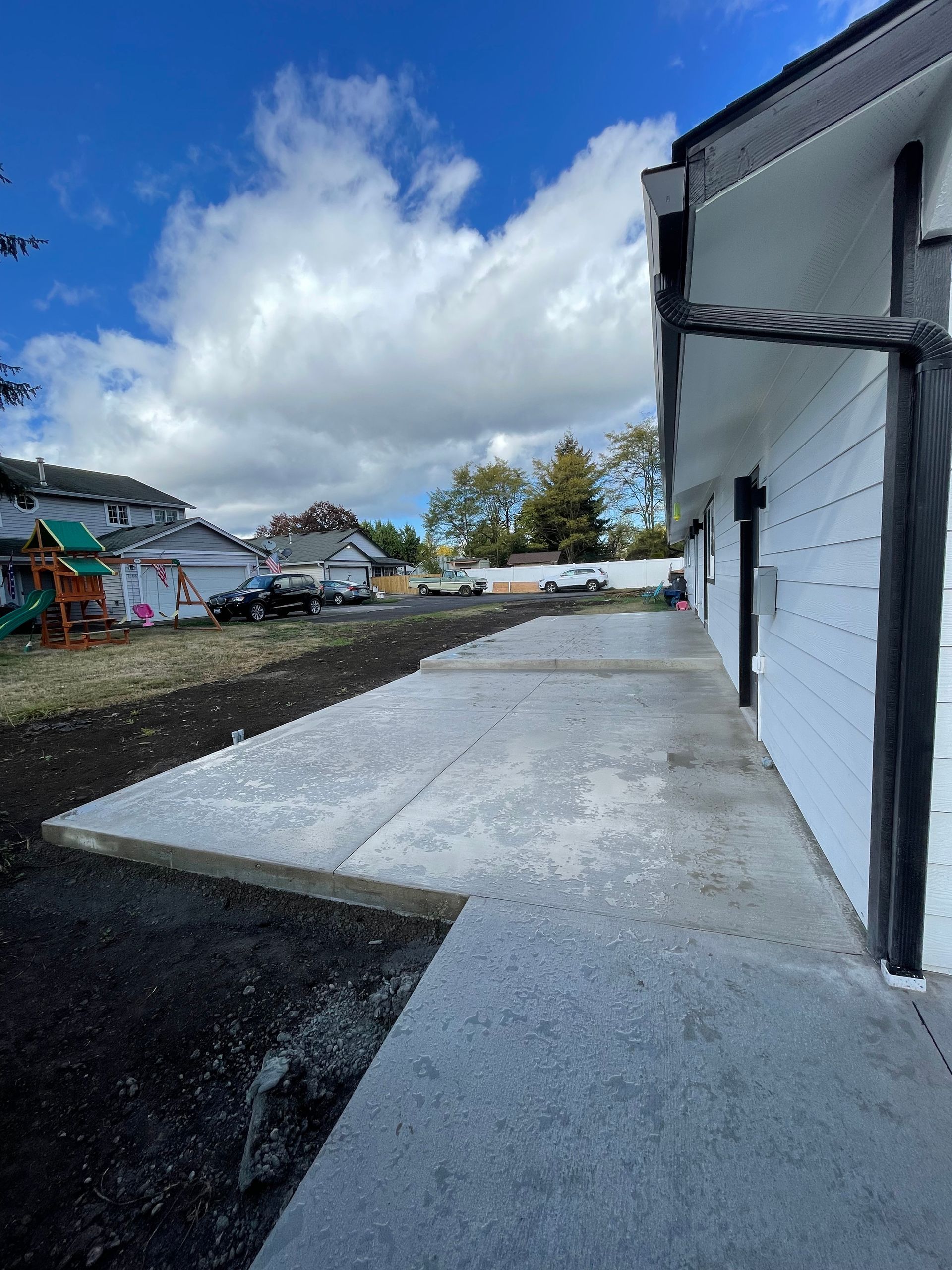 A newly poured concrete walkway runs along the side of a white house, bordered by bare dirt and a residential backyard.