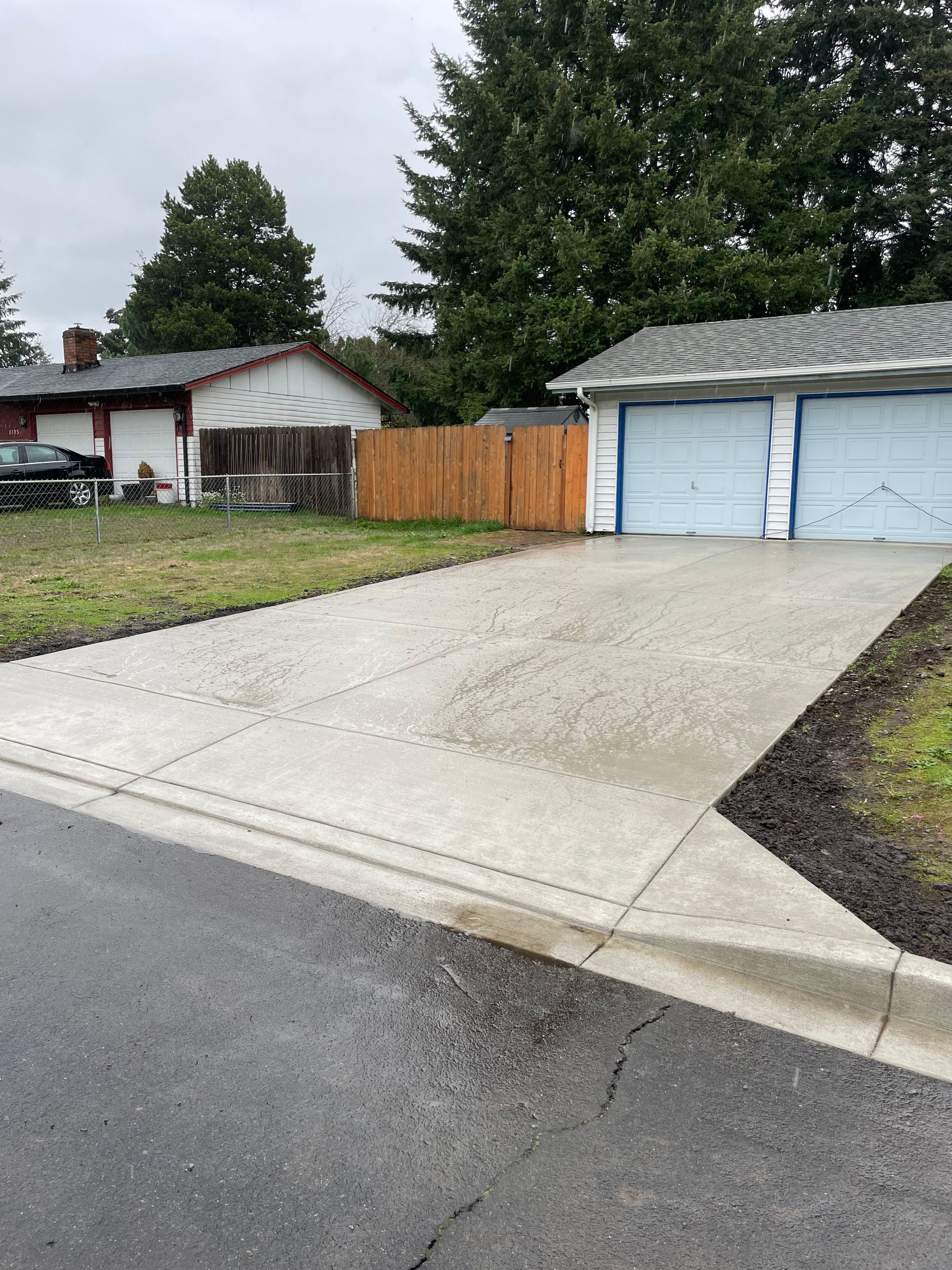 A newly poured concrete driveway leads to a two-car garage next to a wooden fence and a grassy yard.