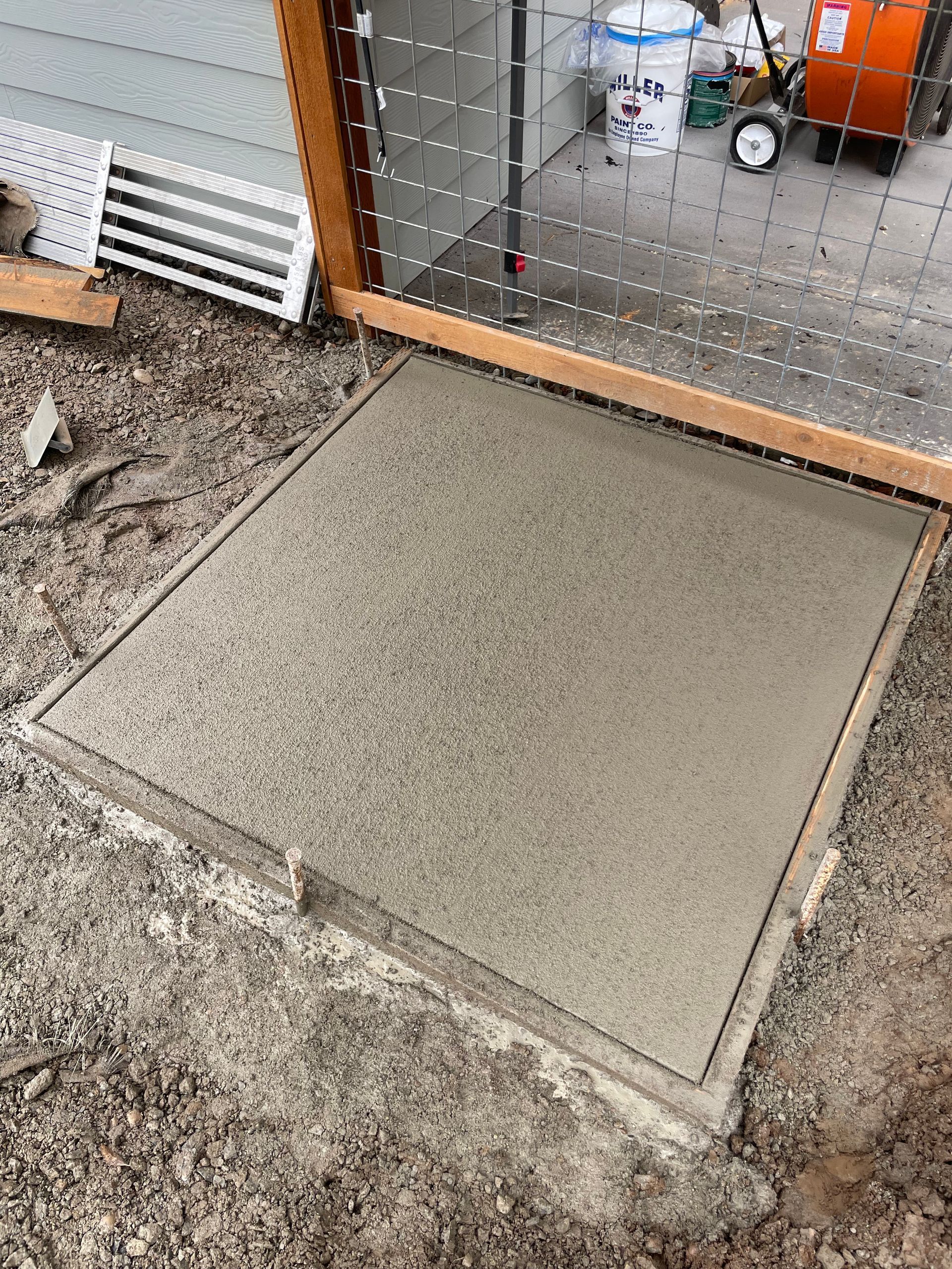 A newly poured square concrete slab sits on the ground in front of an outdoor kennel area.
