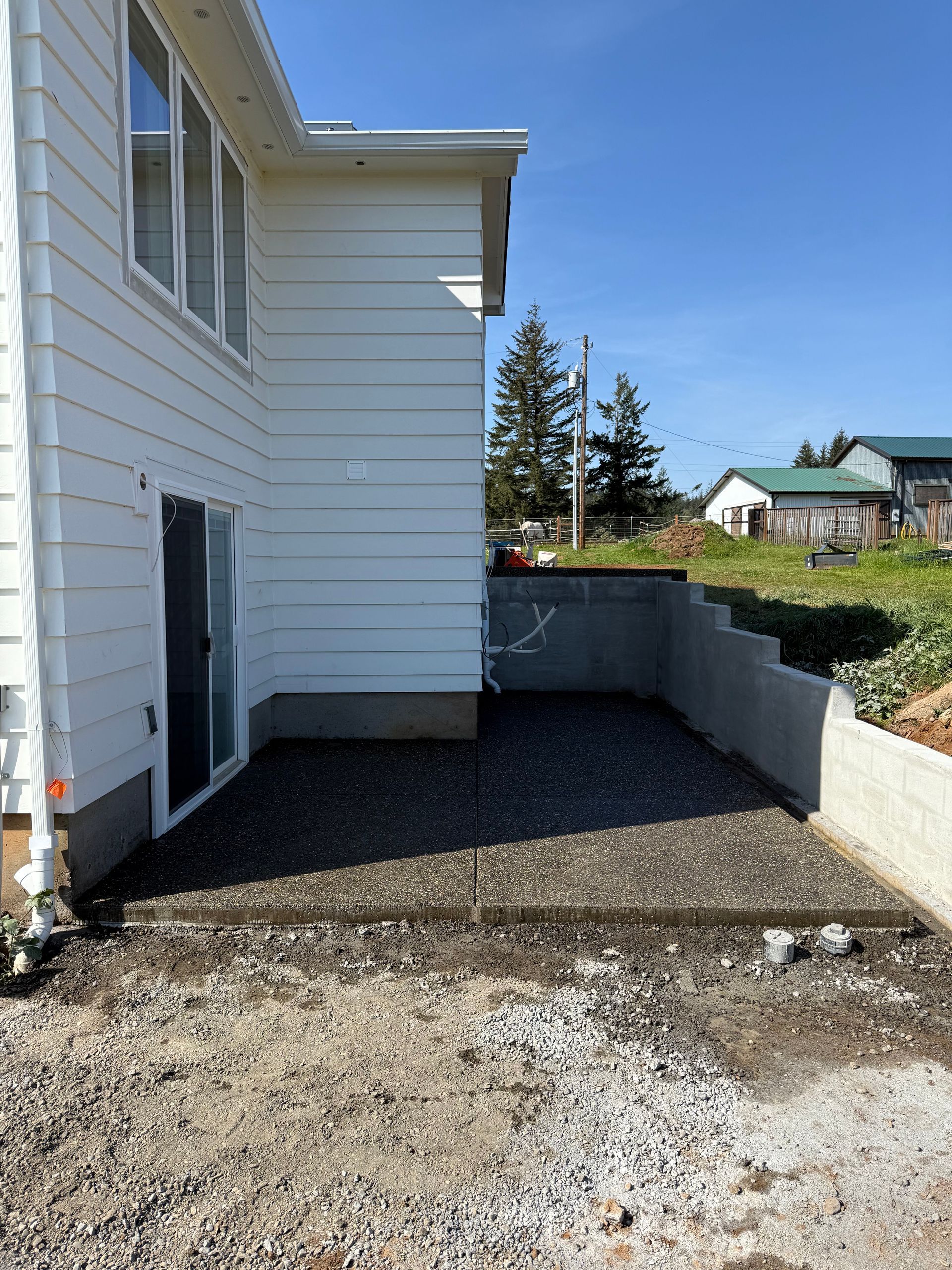 A newly poured concrete patio alongside a white house with horizontal siding and a retaining wall in a backyard setting.