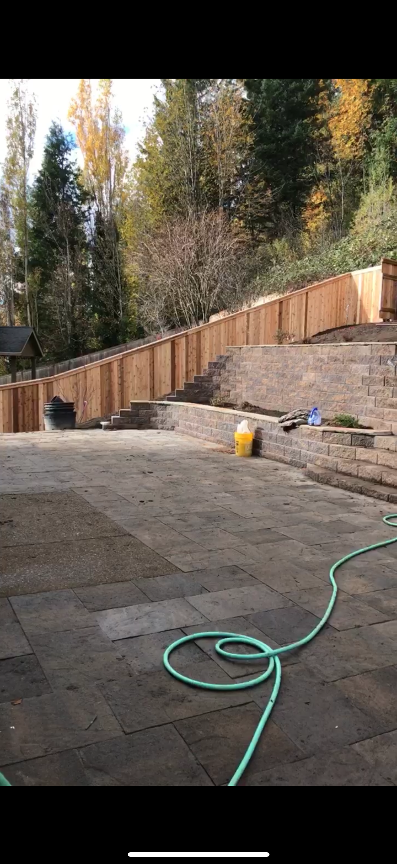A patio with paved stones, a concrete retaining wall, and a new wooden fence sloping up a hill with trees in the background.