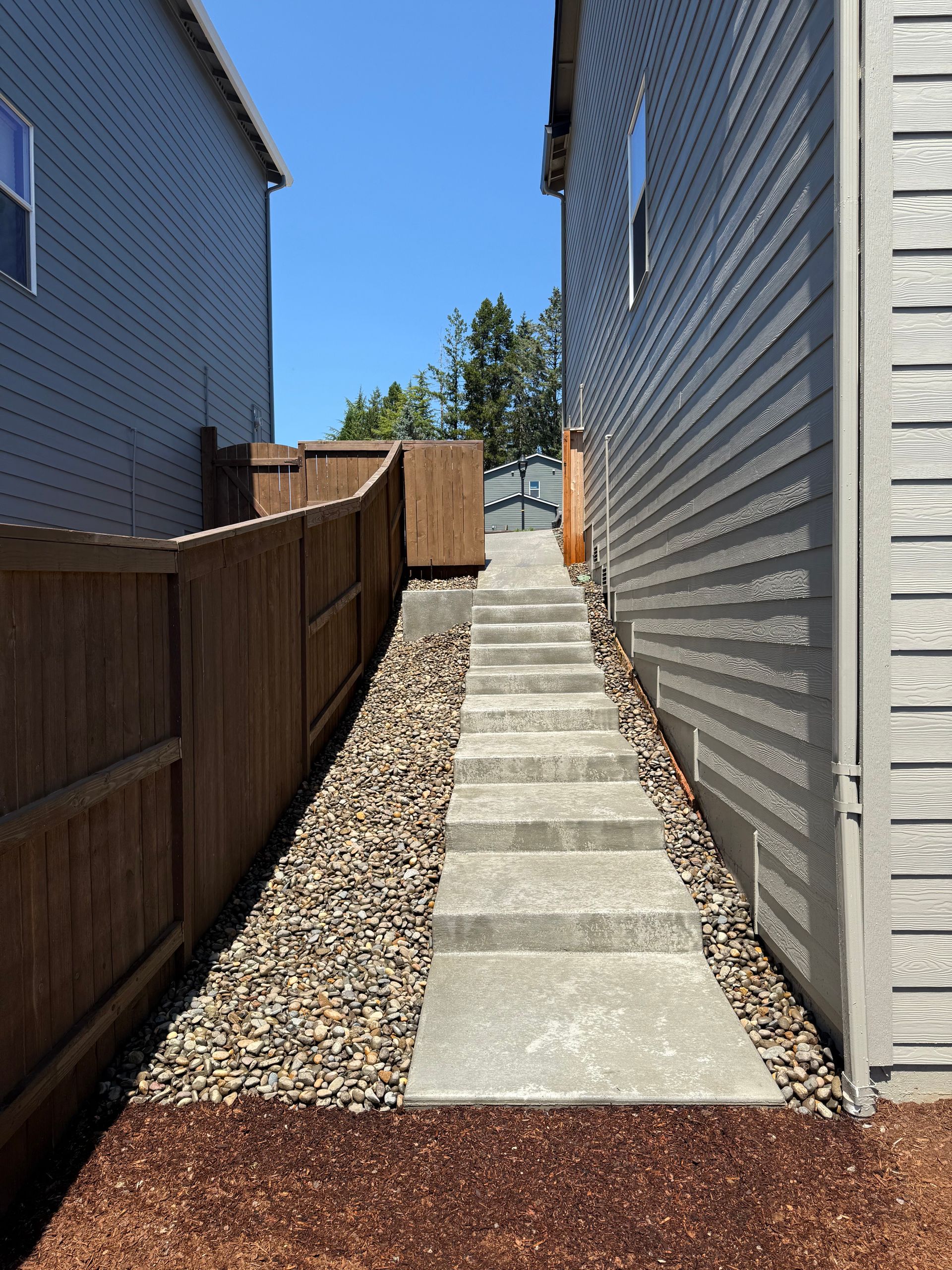 A narrow concrete staircase leads up between two houses, flanked by a wooden fence on one side and gravel on the other.