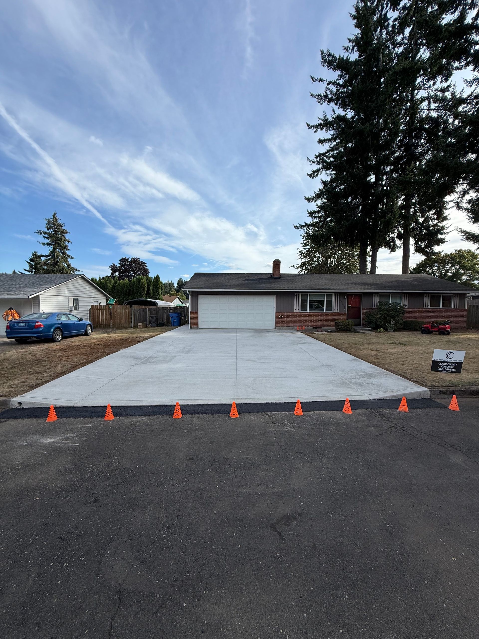 A freshly poured concrete driveway leading to a suburban ranch-style house, marked by a line of orange traffic cones.