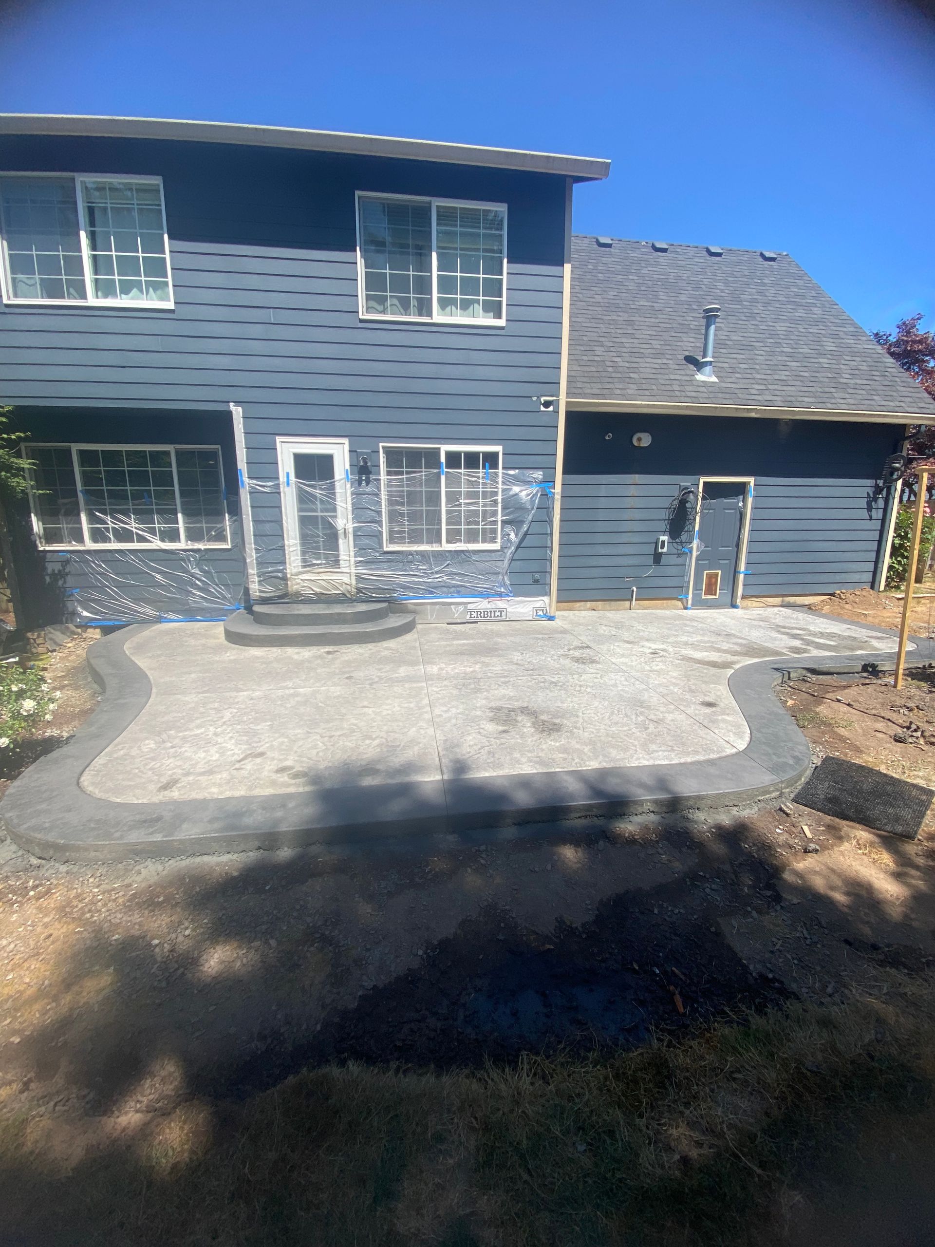 A blue two-story house with a newly installed grey stone patio and curved border under a clear blue sky.