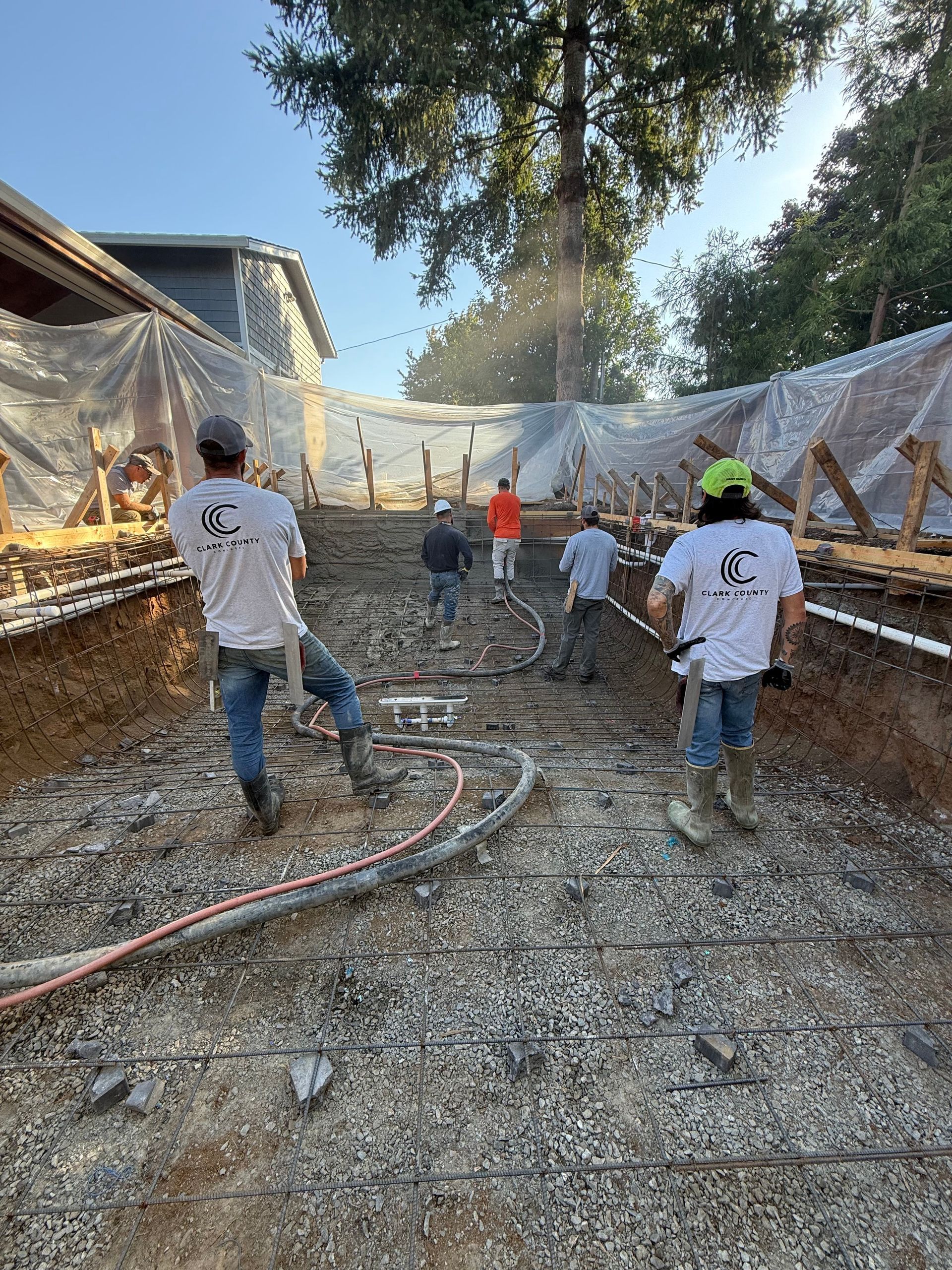 A crew prepares a construction site with rebar and gravel inside a pool-shaped excavation area lined with plastic sheeting.