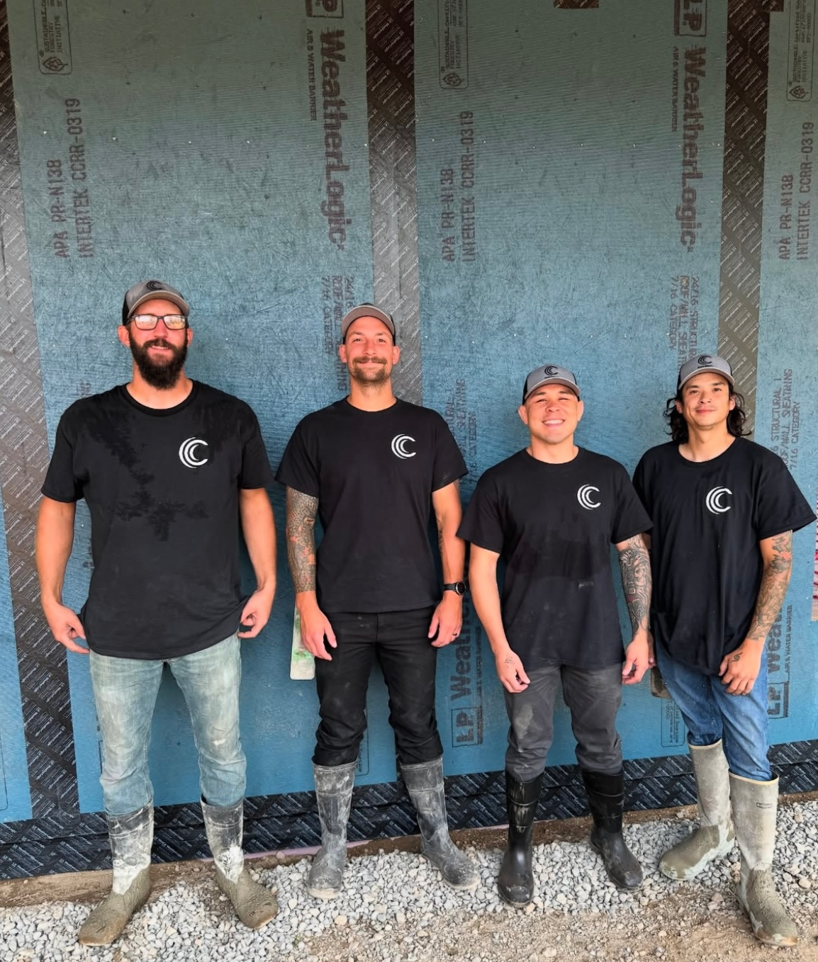 Four people in matching black t-shirts and work boots stand side-by-side in front of a blue structural wall covering.