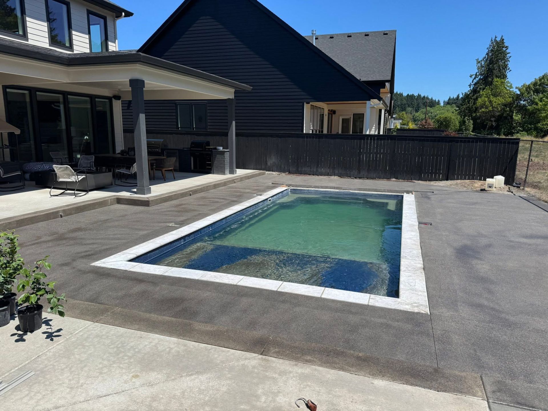A rectangular pool with a white stone border sits in a backyard next to a house with a covered patio.