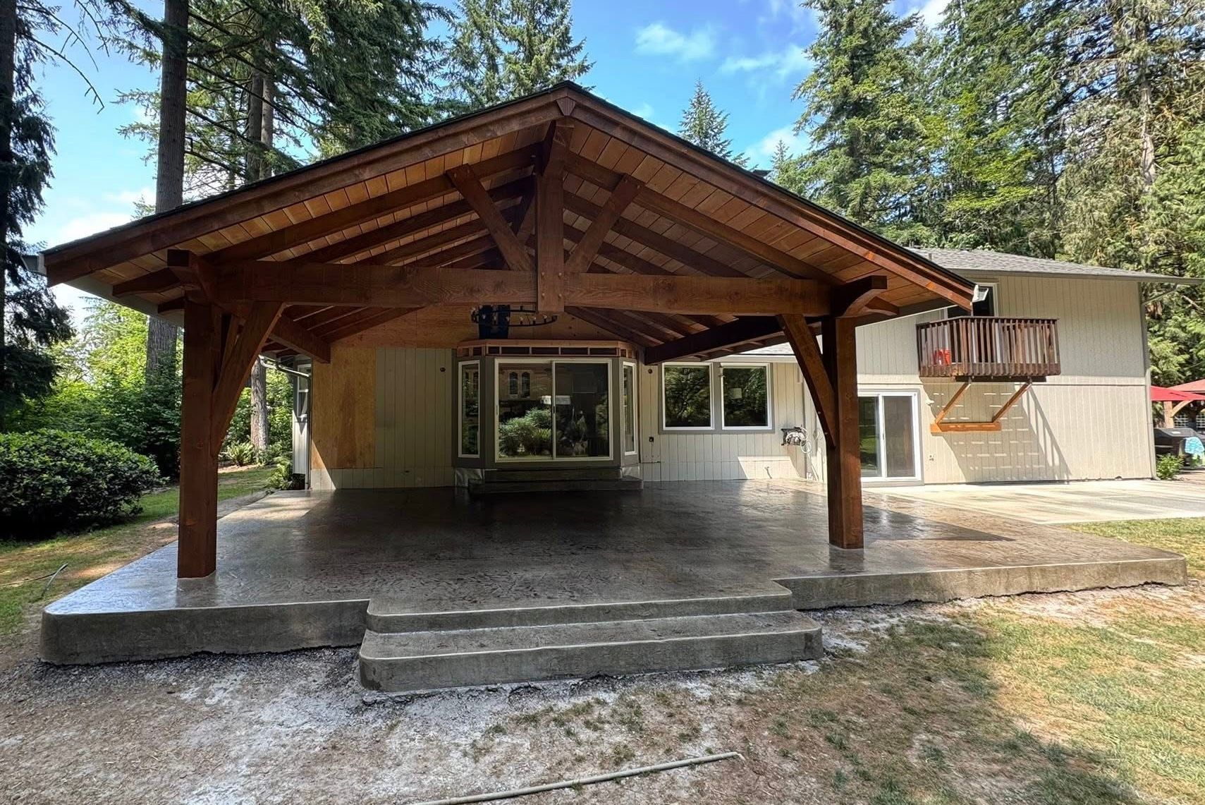 A large wooden patio cover with a gabled roof stands over a concrete patio outside a house with beige siding and trees.