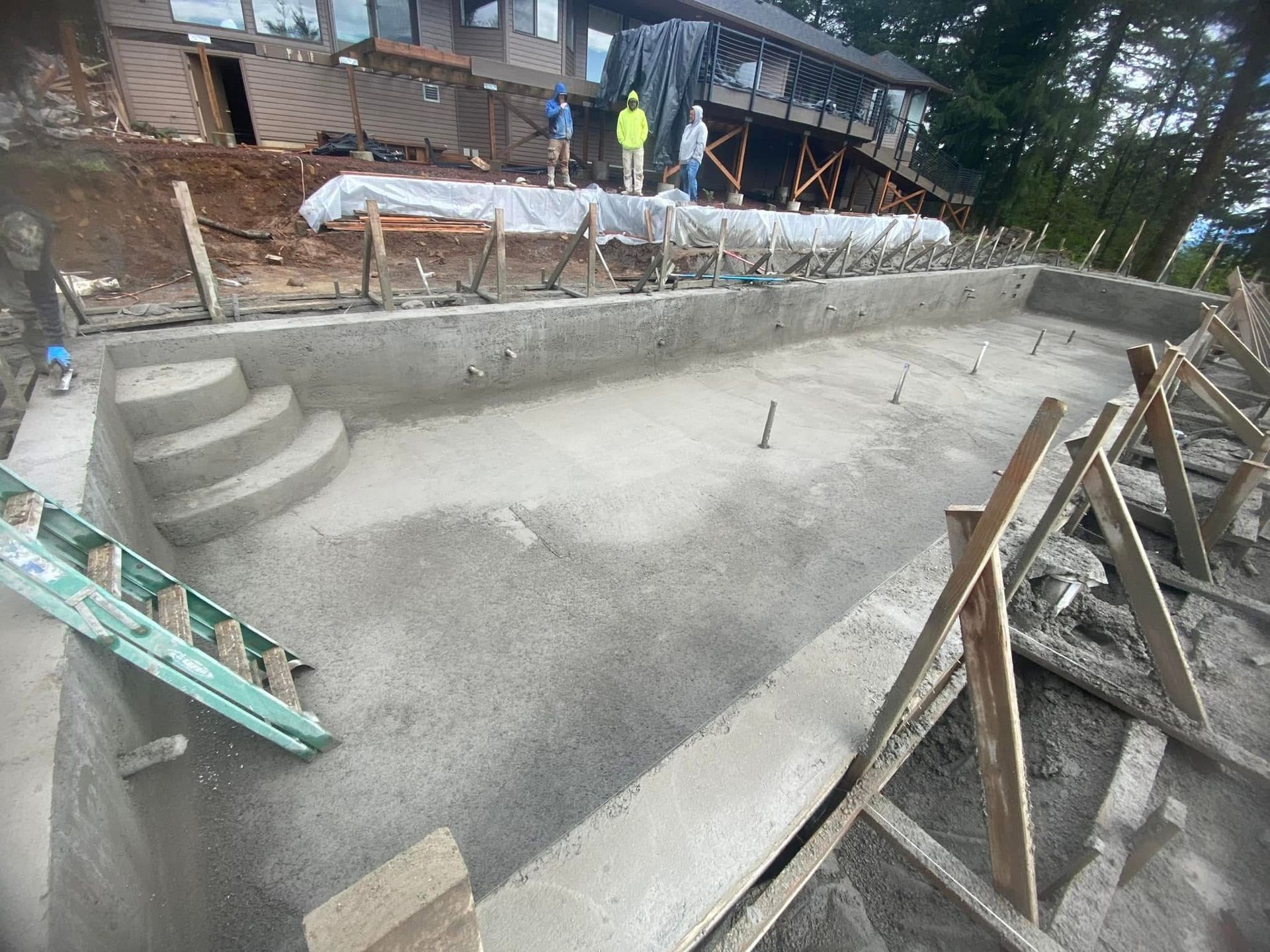 A concrete swimming pool under construction with wooden framing, a ladder, and workers in the background.