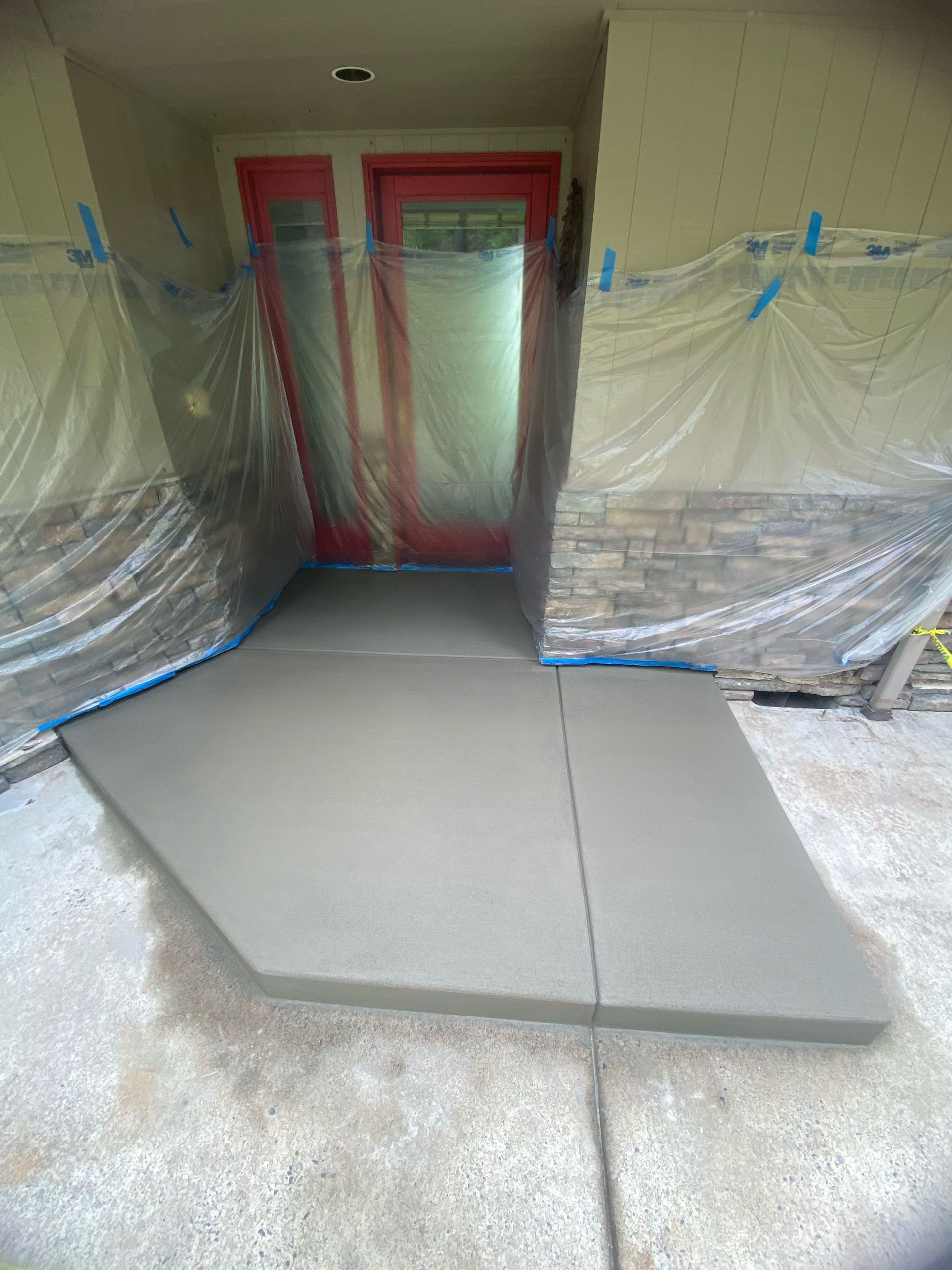 A newly poured concrete step leads to a red door, with plastic sheeting covering the entryway and surrounding stone walls.