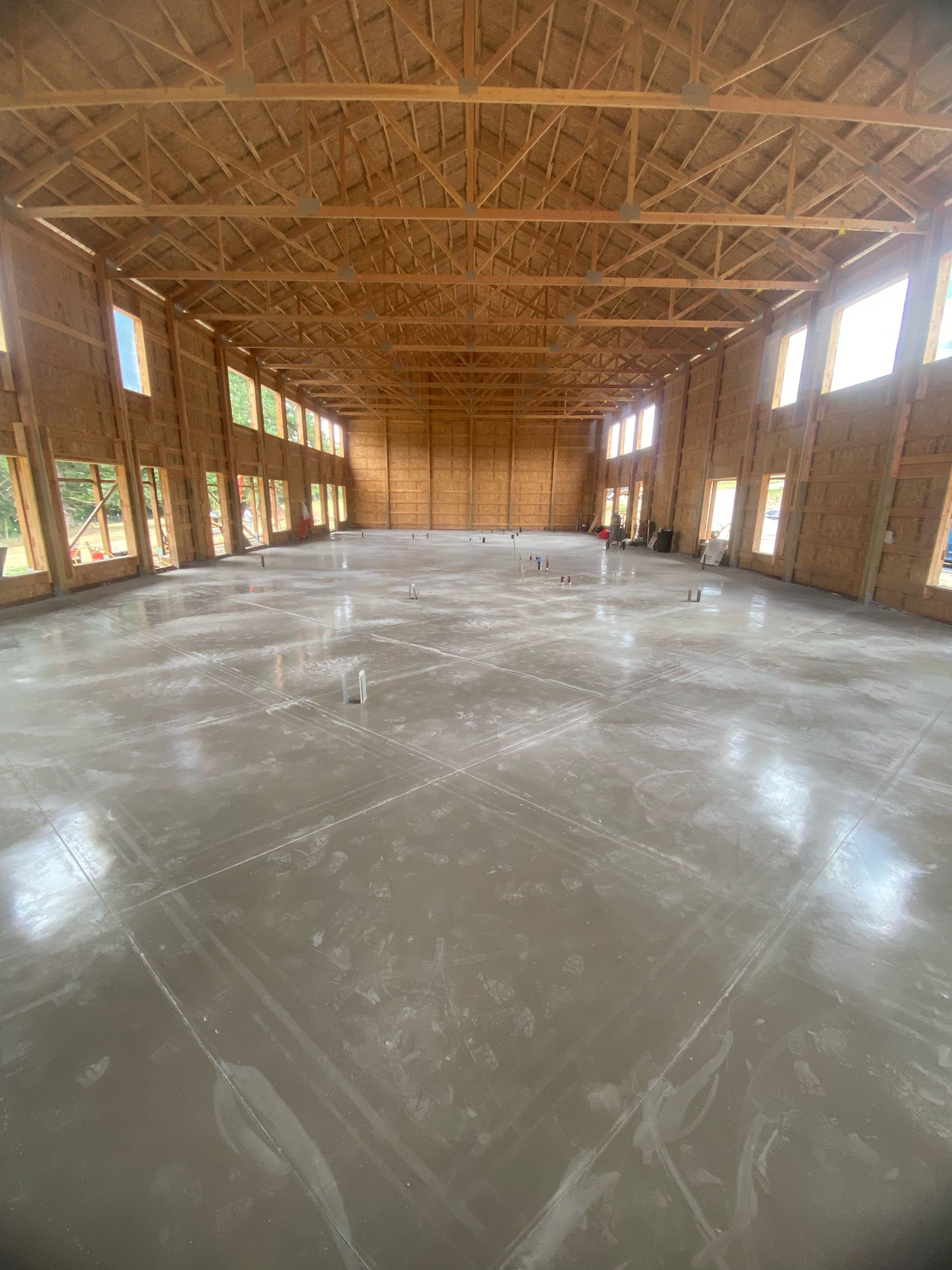 Interior of a large unfinished building with wooden wall framing, exposed roof trusses, and a smooth concrete floor.