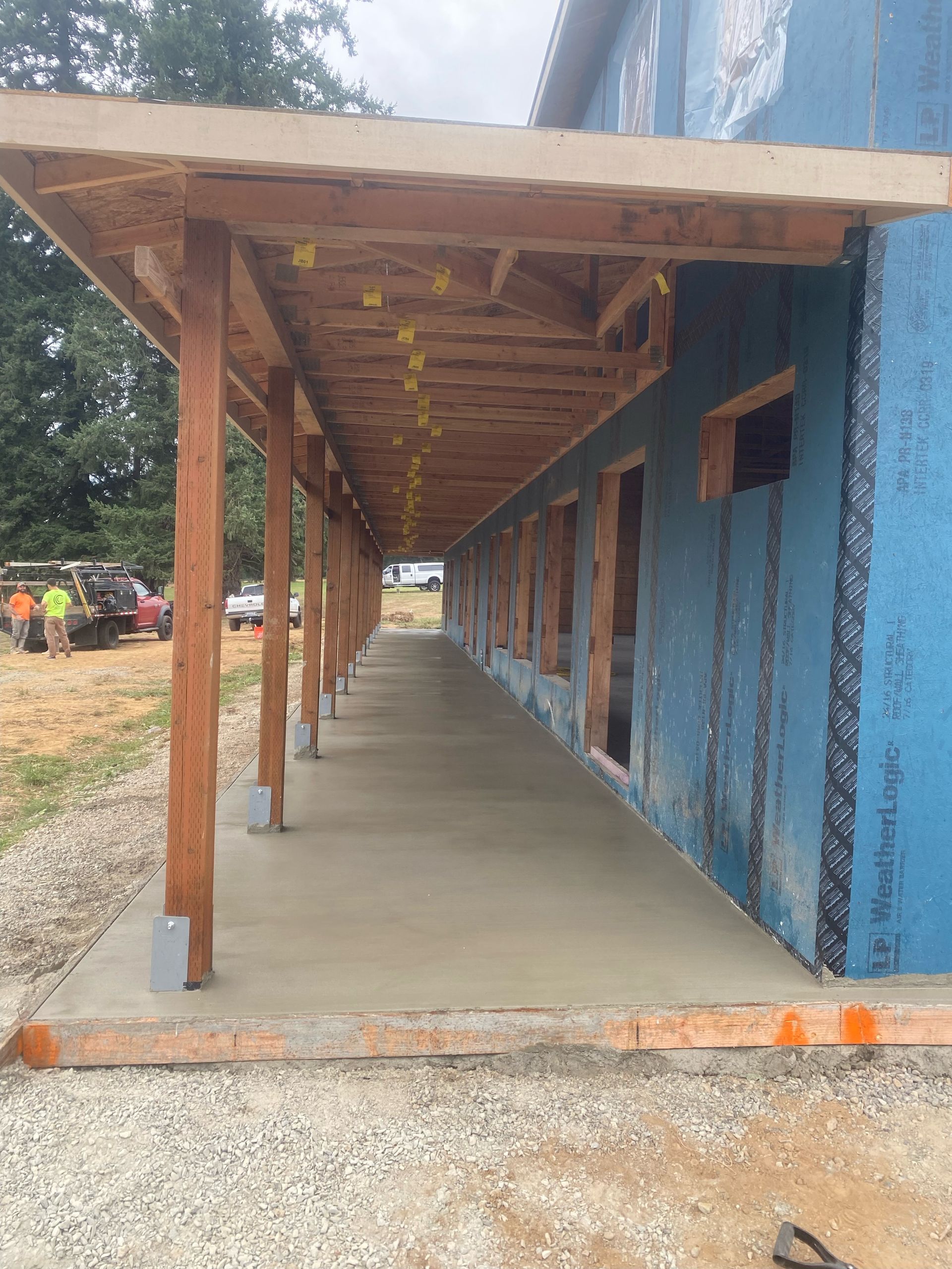 A covered porch under construction runs along the side of a building with blue exterior sheathing.