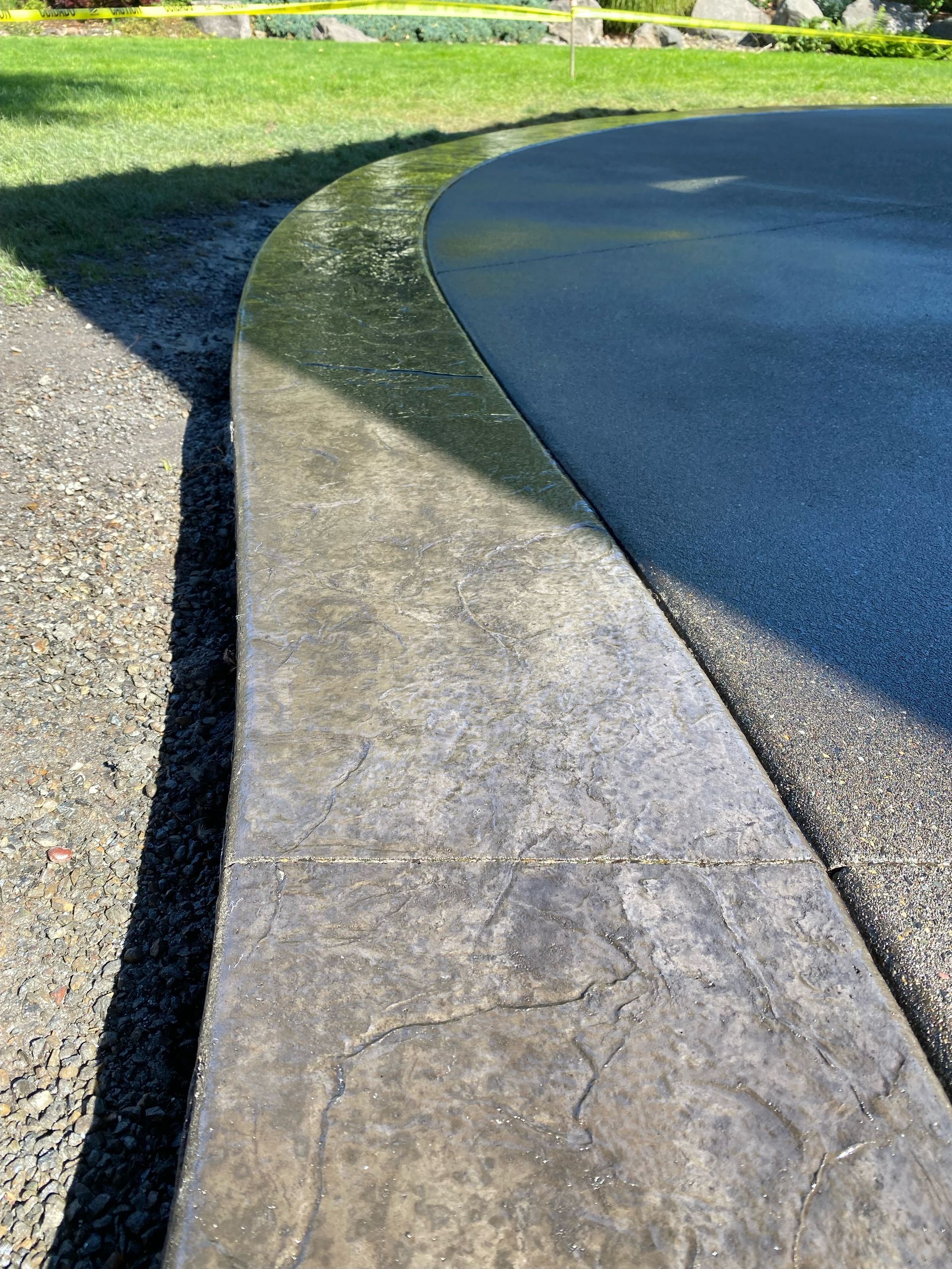 A curved, textured concrete border separates a dark paved area from a gravel path and grassy lawn.