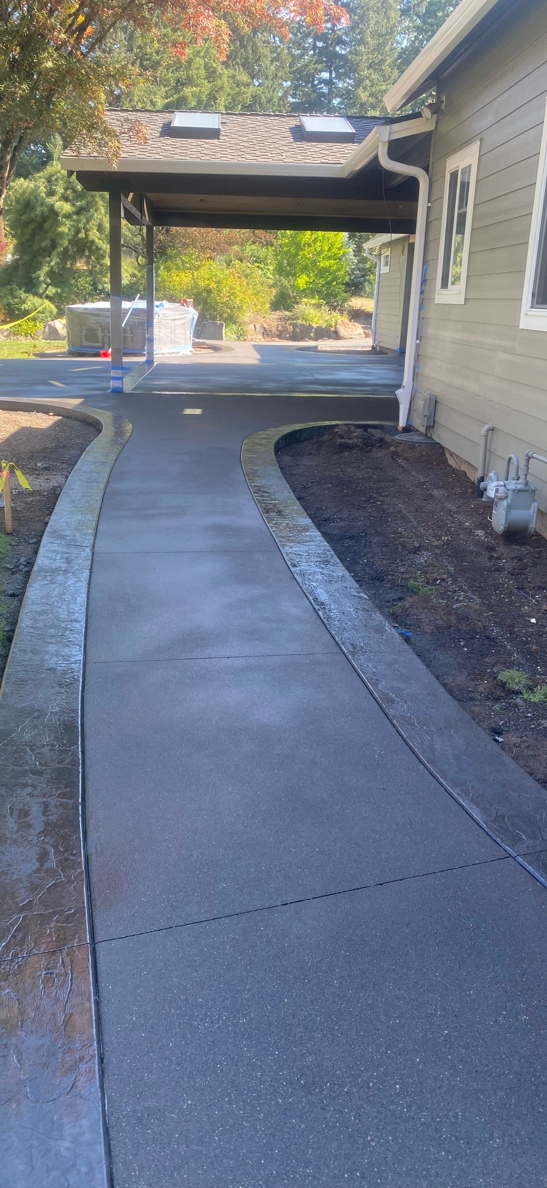 A concrete pathway leads toward a covered carport area next to a light-colored building with dark mulch borders.