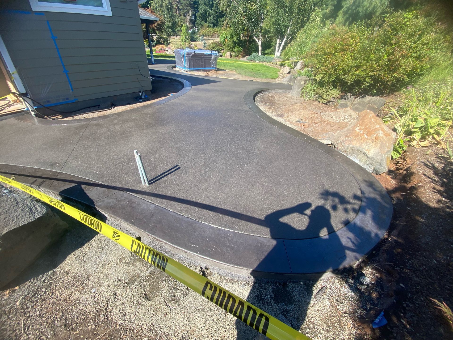 A newly paved, curved dark asphalt walkway runs alongside a house, cordoned off by yellow caution tape.