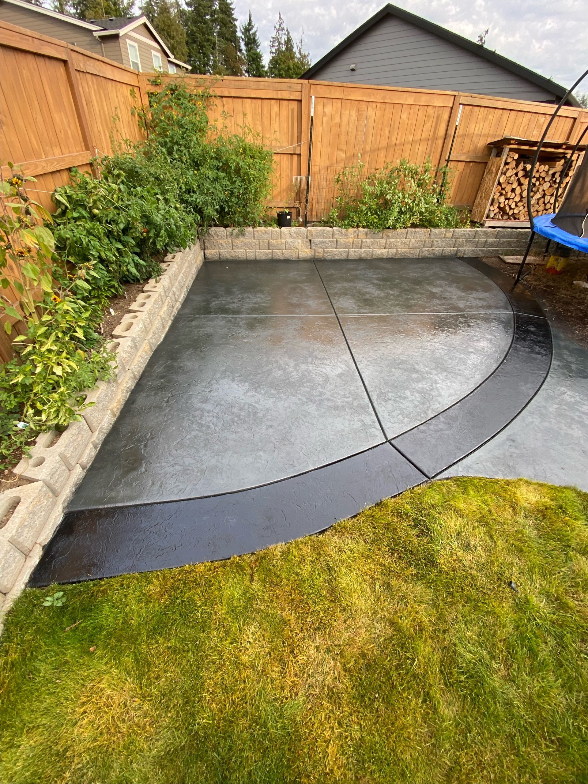 A backyard patio featuring a semi-circular dark concrete design bordered by a raised stone garden bed and lush greenery.