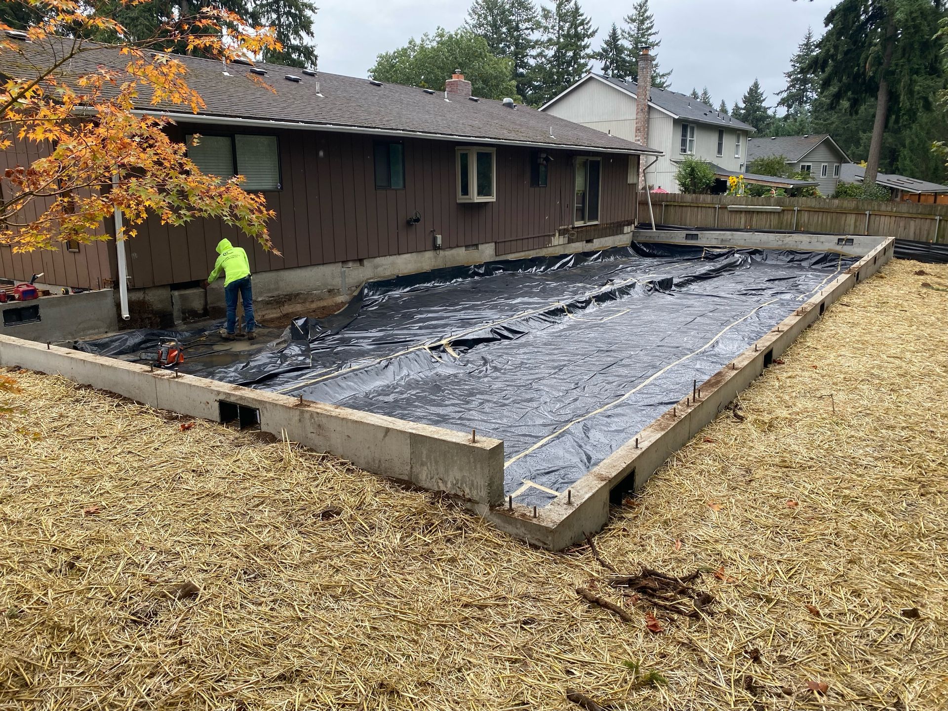 A construction worker in a high-visibility jacket stands beside a rectangular concrete foundation filled with plastic.