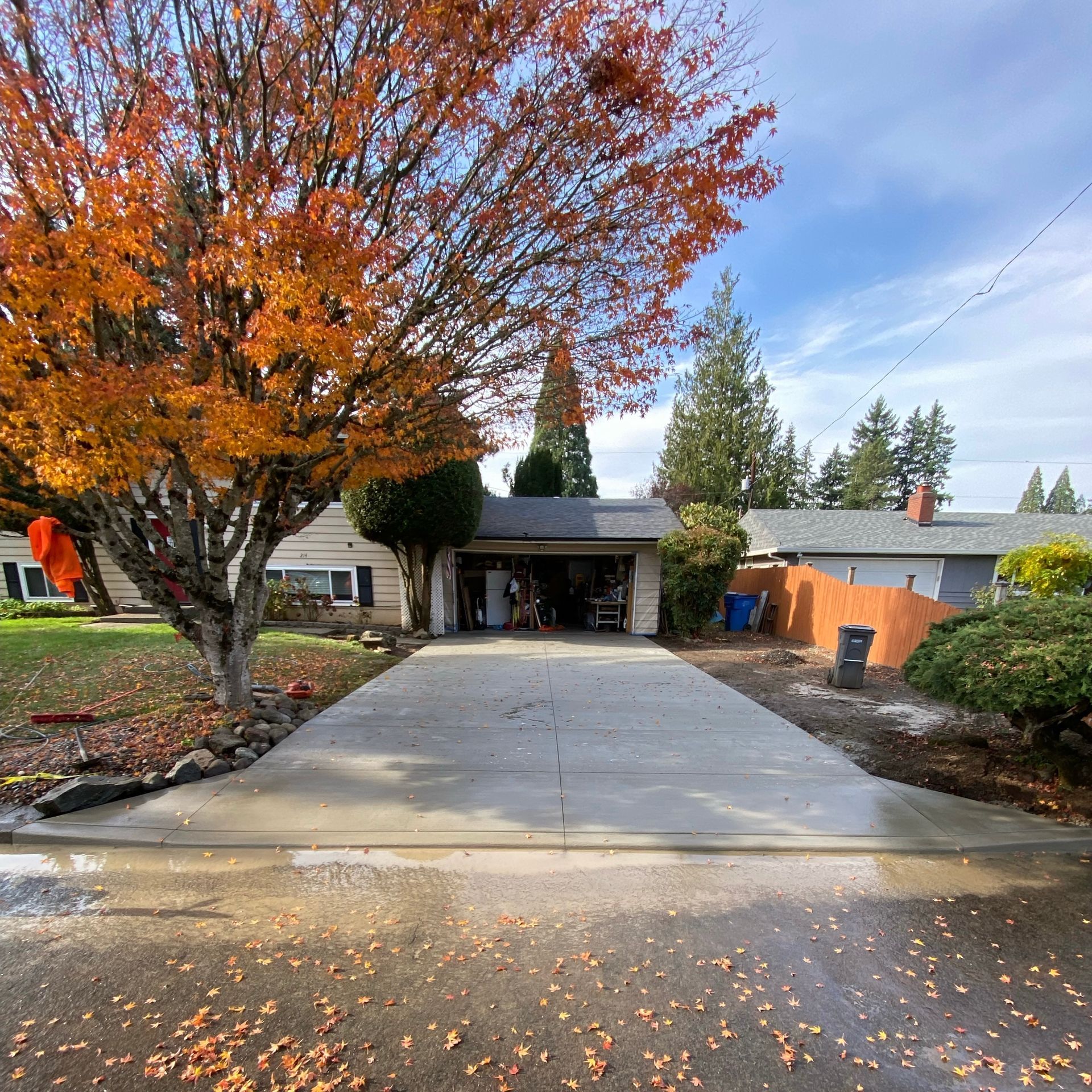 A newly poured concrete driveway leads to a suburban home with a garage, flanked by a vibrant orange autumn tree and lawn.