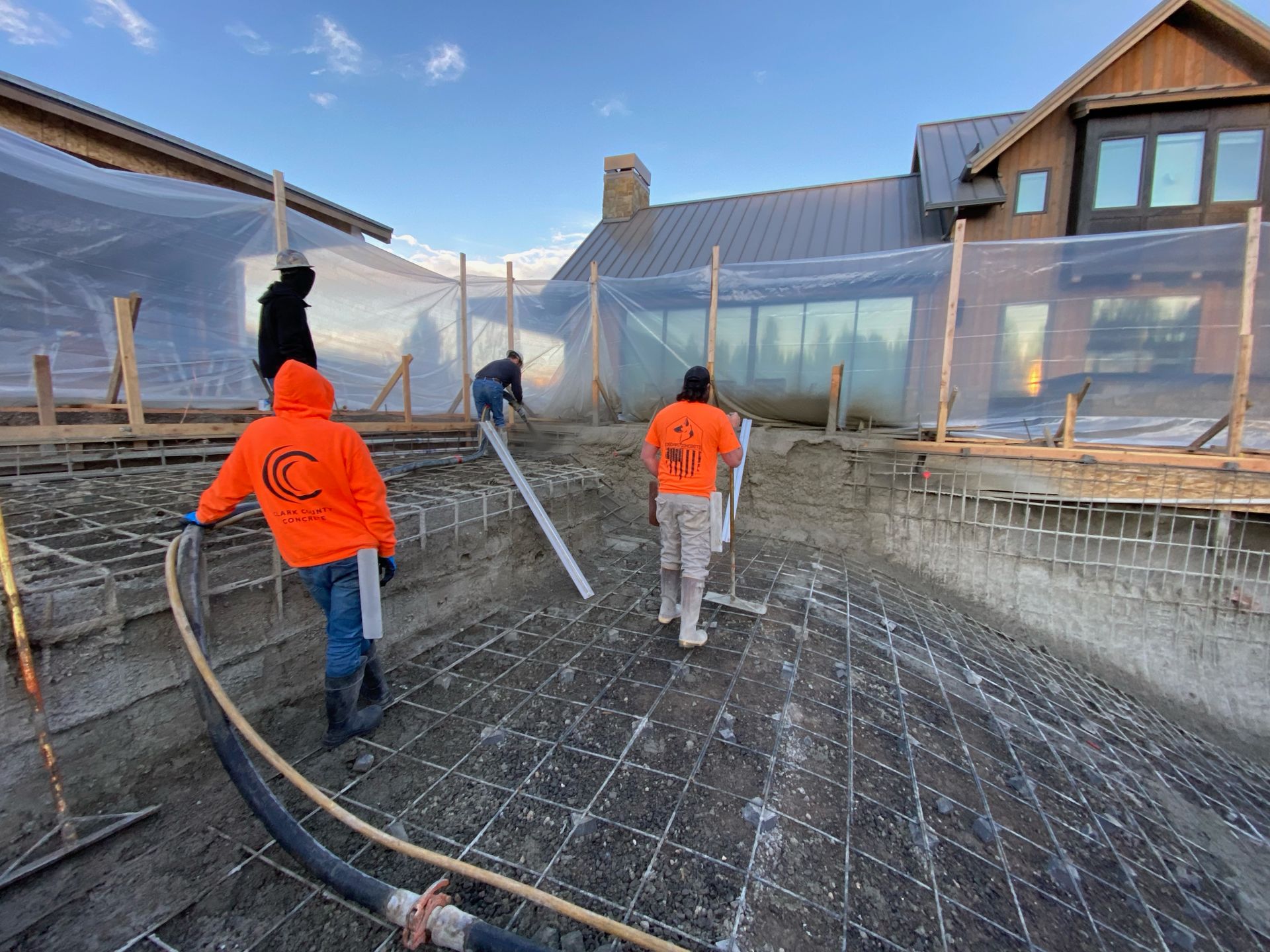 Construction crew working on a concrete pool foundation outside a large house surrounded by protective plastic sheeting.
