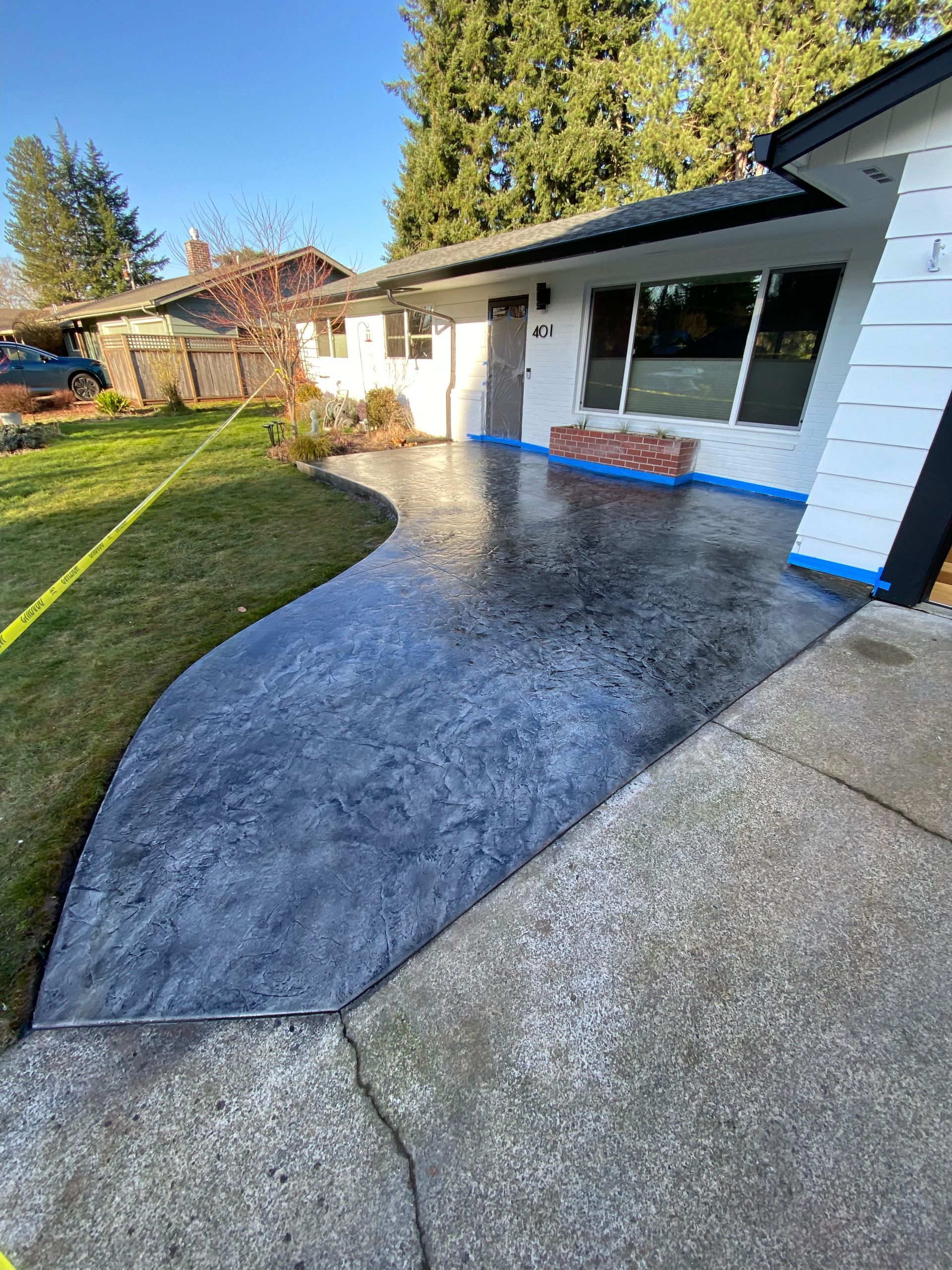 A newly stained, textured dark blue patio area curves against a white house and an older concrete driveway.