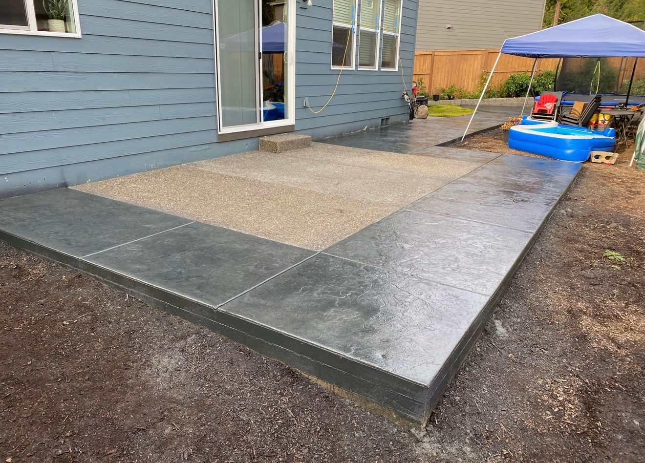 A newly poured concrete patio with a textured light-brown center bordered by smooth dark-gray slabs next to a blue house.