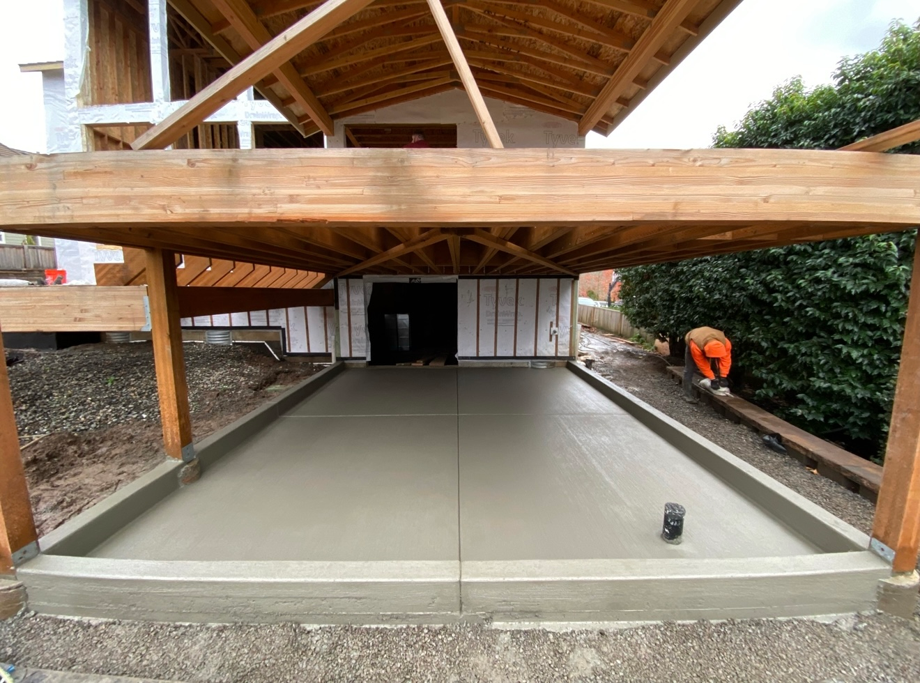 Newly poured concrete slab under a wooden deck structure with a person in orange safety gear working nearby.