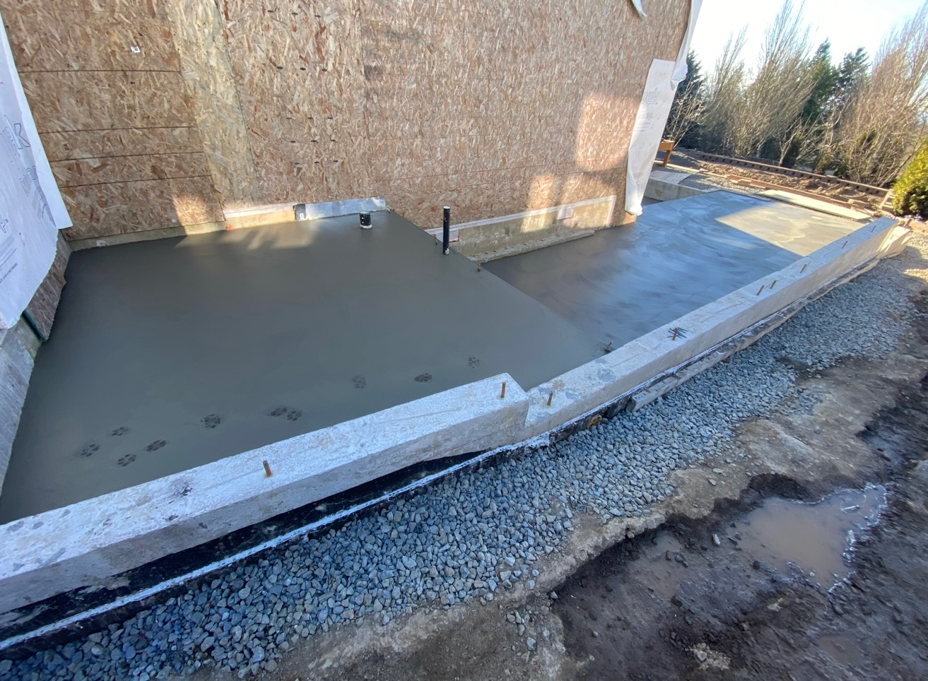 Freshly poured concrete slab and foundation walls along the side of a wooden building under construction.