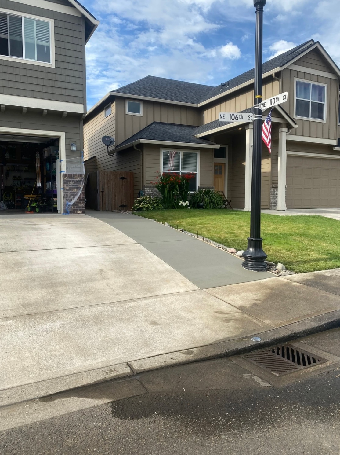 A multi-story suburban house with a concrete driveway and sidewalk, featuring an American flag near a street lamp.