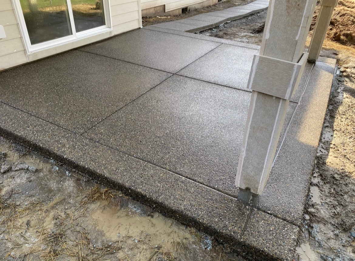 A freshly poured gray concrete patio with a textured aggregate finish, situated next to a house with a sliding door.
