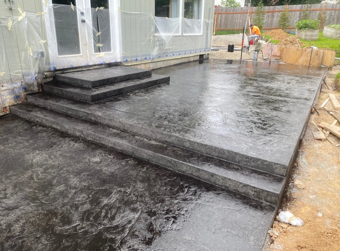 Newly poured dark concrete patio with two levels of steps leading up to French doors on a house under construction.