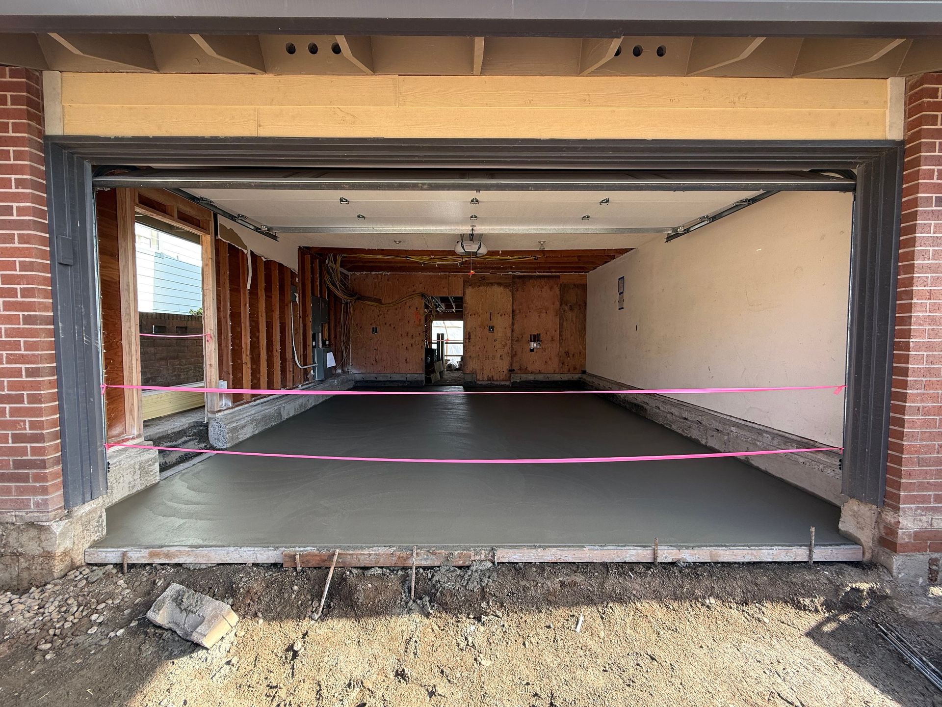 A newly poured concrete floor inside an unfinished garage framed with brick, with a pink caution tape barrier.