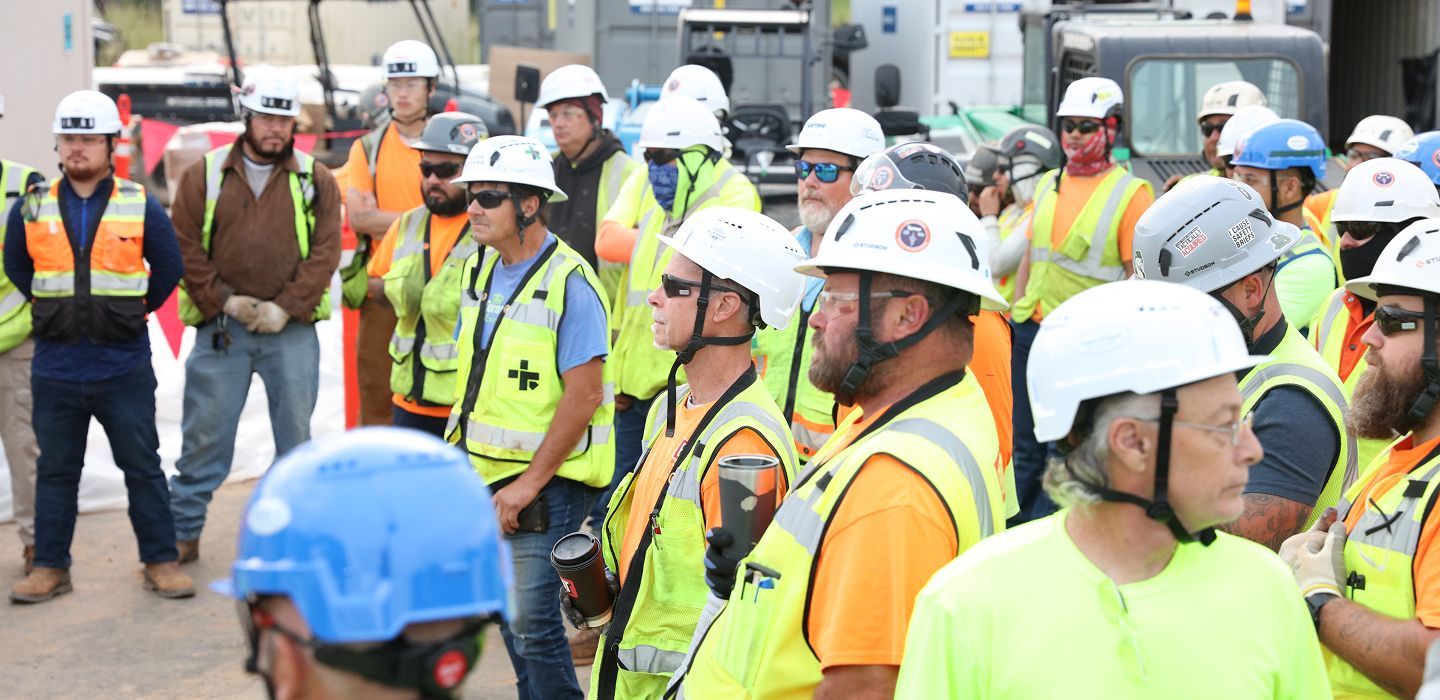 Group of construction workers in safety vests and hard hats gathered at a worksite, listening attentively