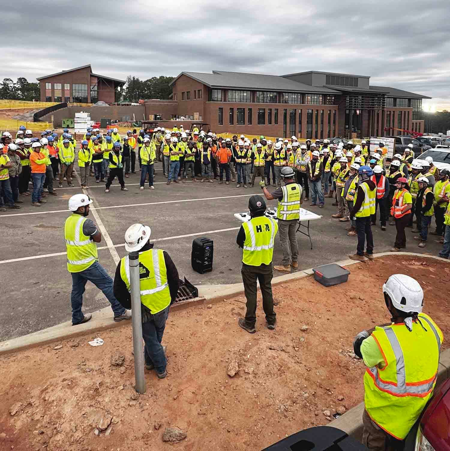 Construction workers in hard hats and safety vests gather at a building site for a safety briefing