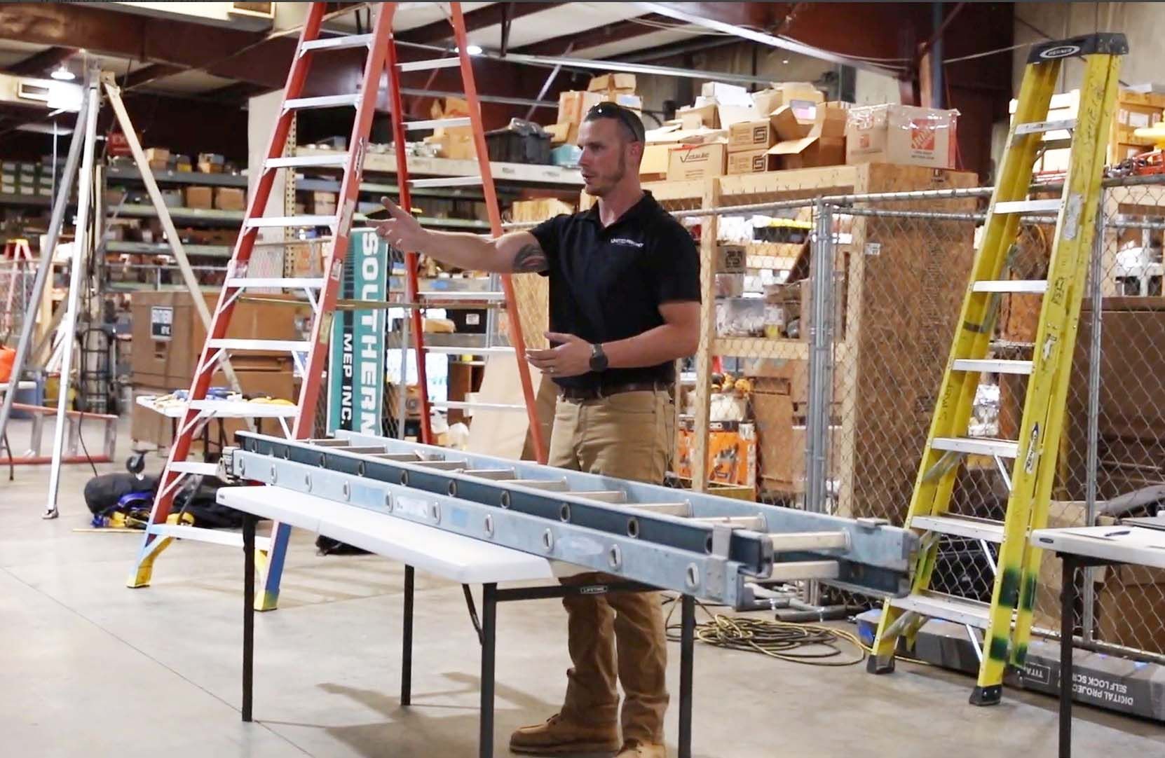 Man standing in a warehouse aisle beside long metal beams and ladders on a worktable