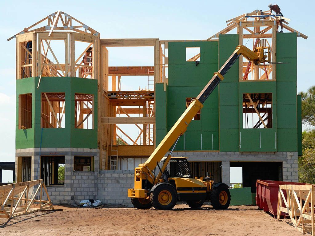 Construction site with two-story wooden building frame, green exterior panels, and a yellow telehandler lifting a beam