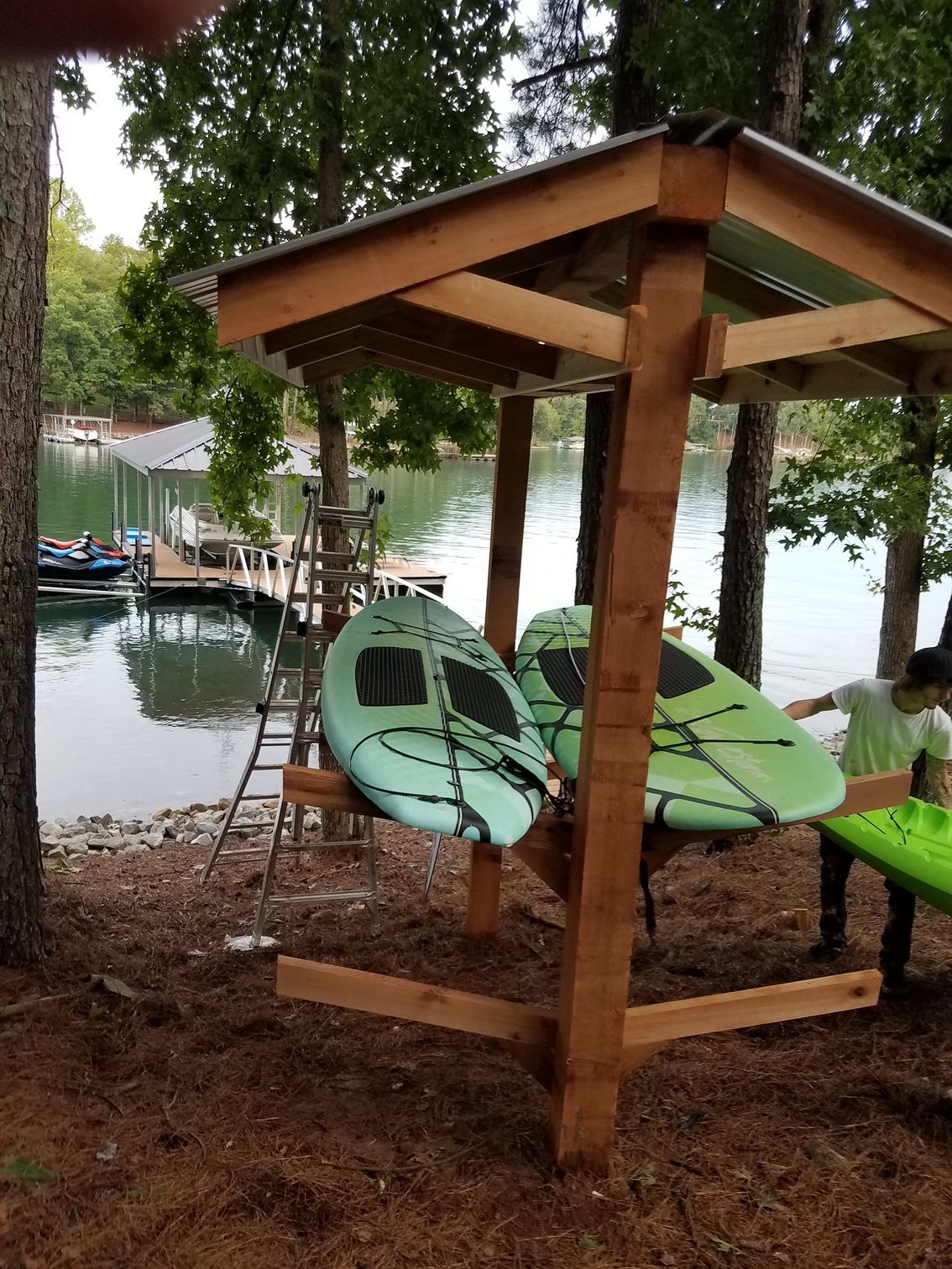Two green kayaks are sitting under a wooden shelter on the shore of a lake.