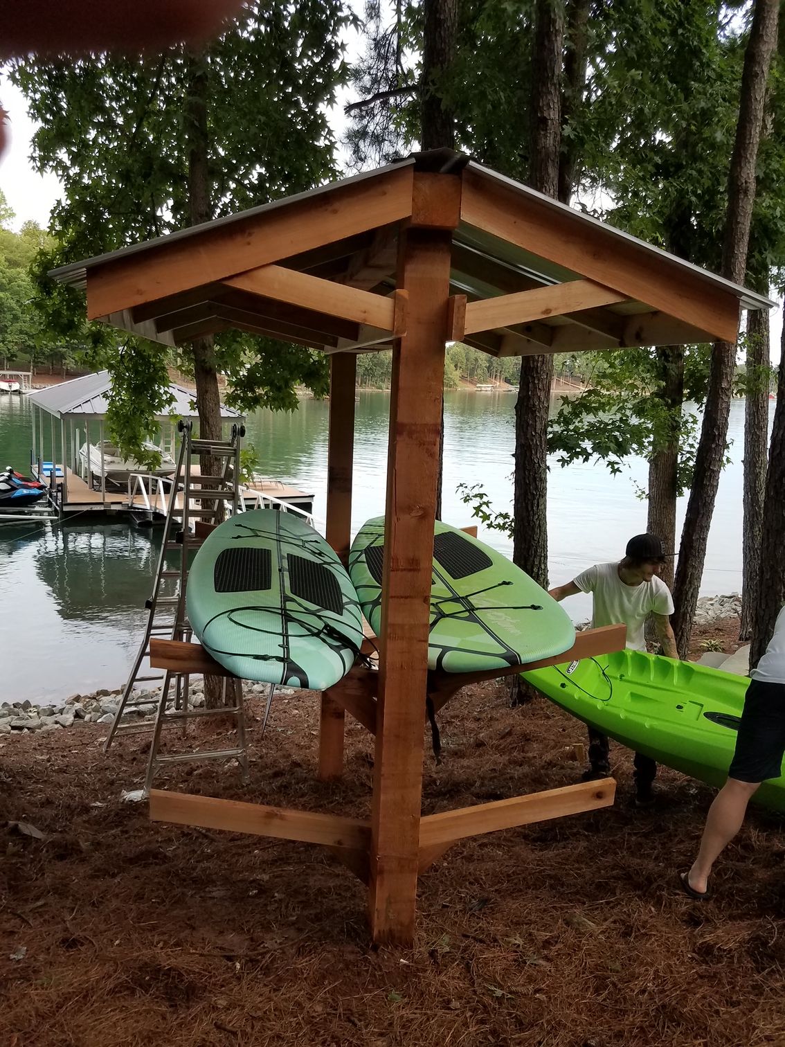 A man is carrying a green kayak under a wooden structure near a lake.