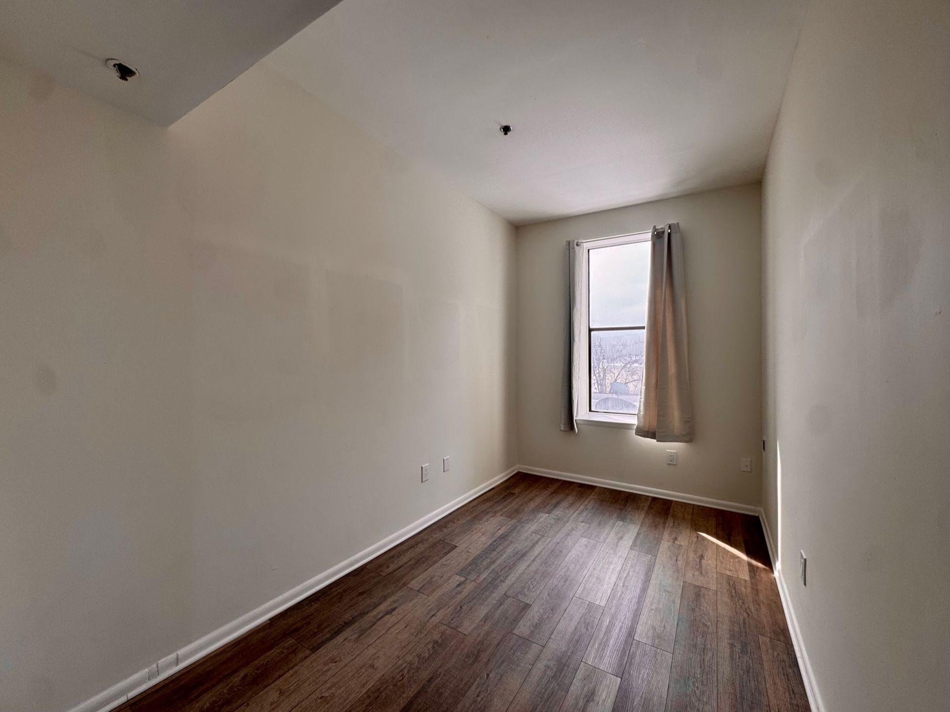 Empty narrow room with white walls, hardwood floor, and a single window with a curtain.