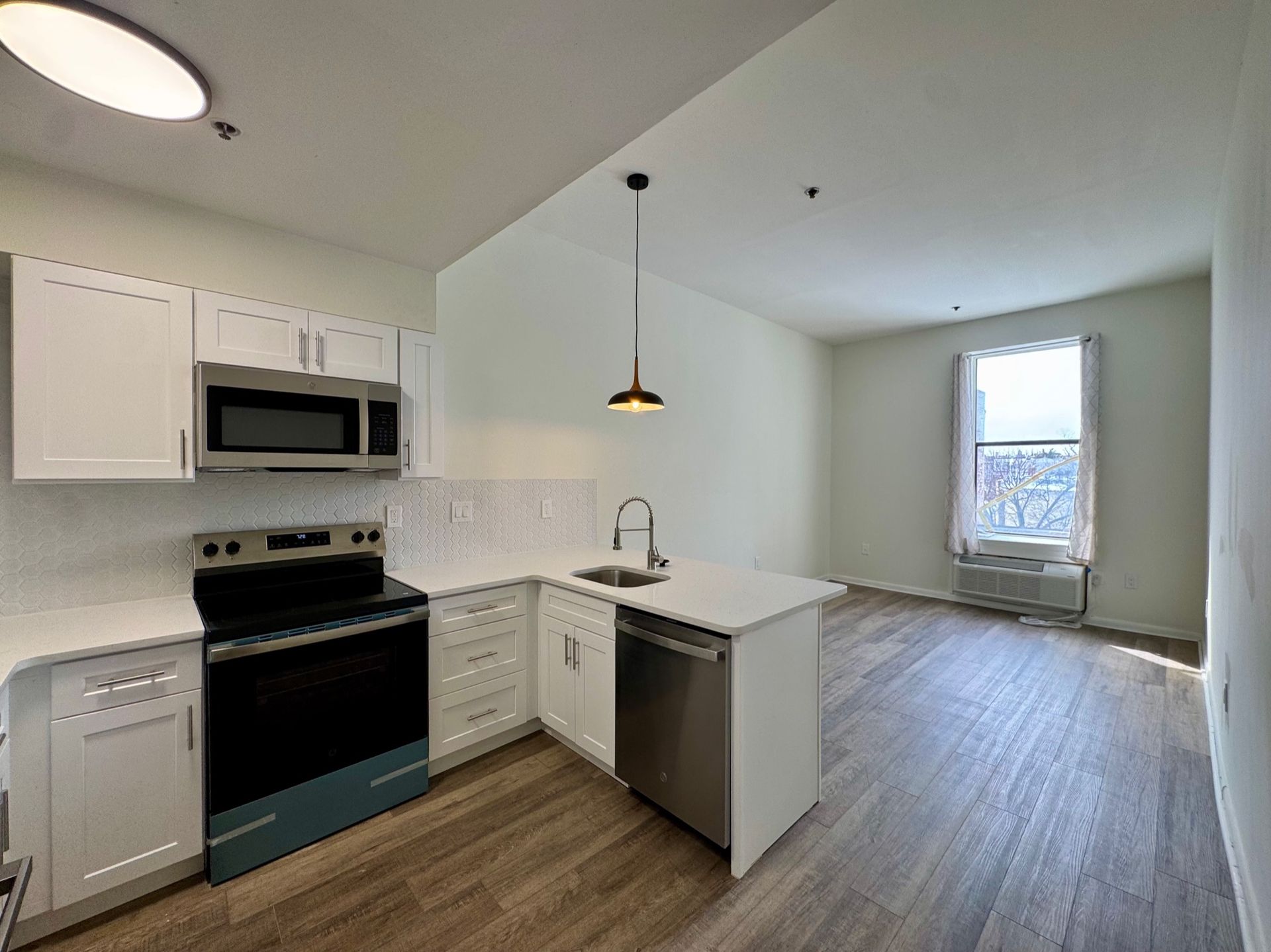 Bright open kitchen with white cabinets, stainless appliances, and hardwood floors, leading to a small living area.