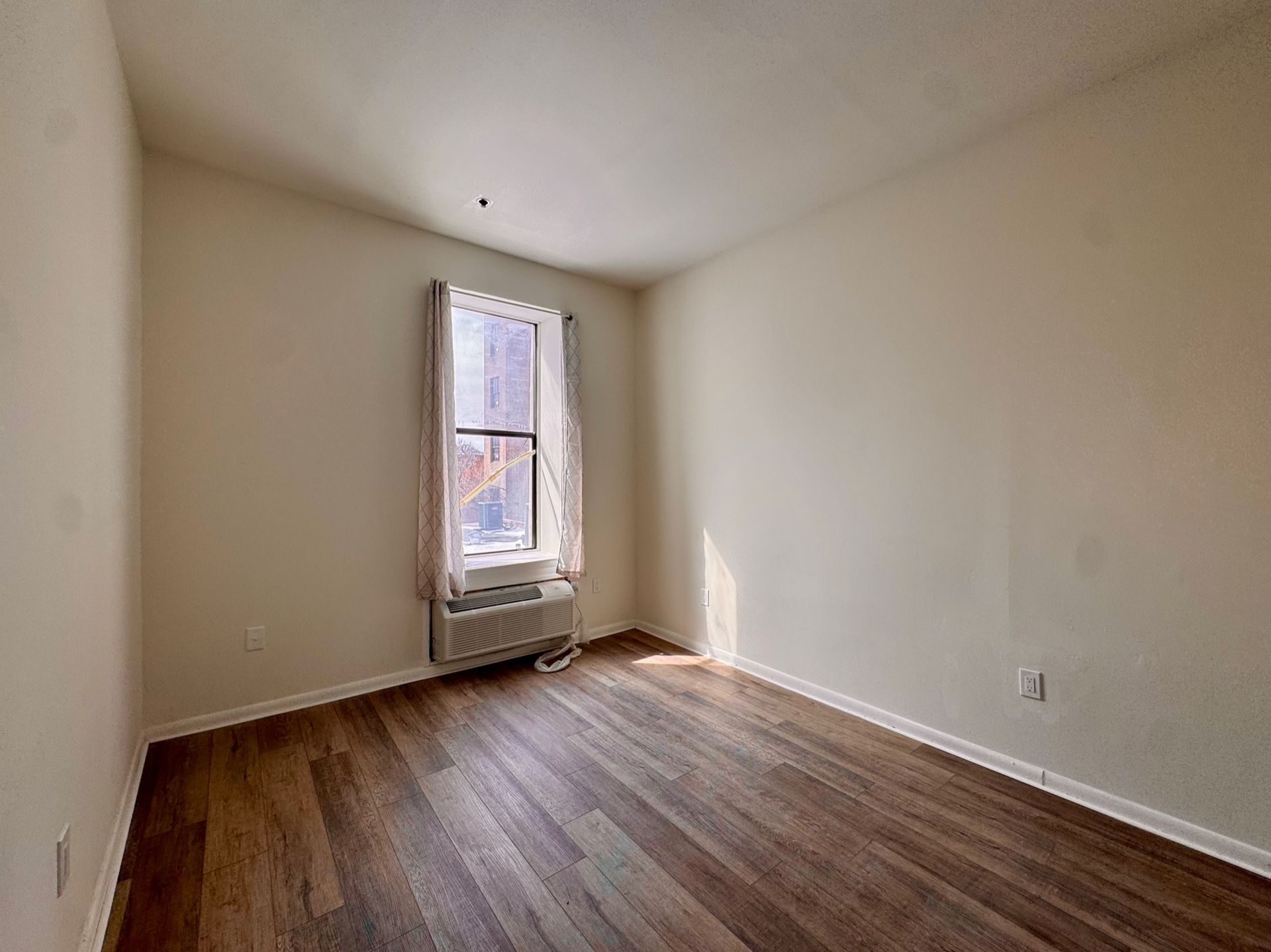 Empty beige room with wood flooring and a single window with a radiator below it